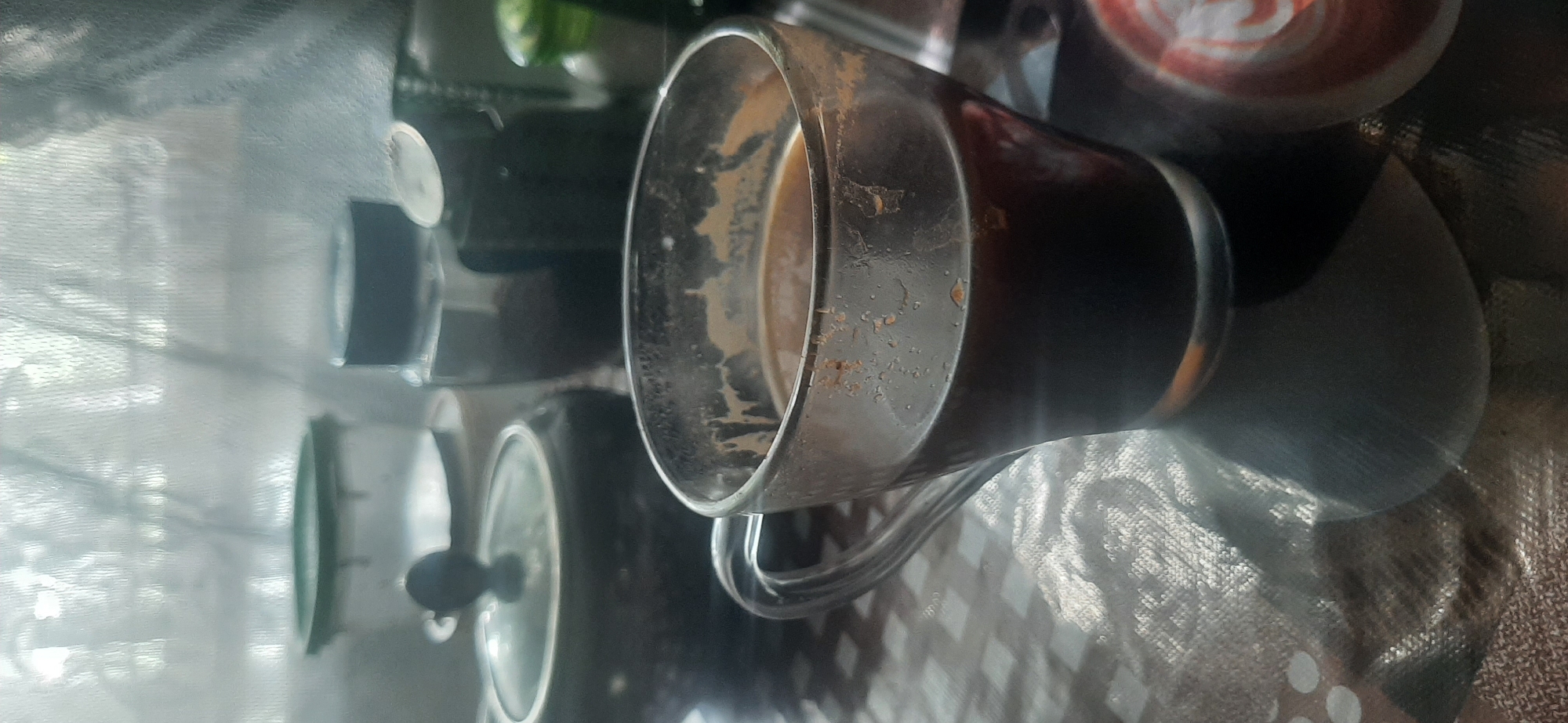 A close-up, slightly blurry, vertical shot captures a glass mug of dark coffee on a checkered tablecloth. The mug has a curved handle and the coffee has visible sediment and some foam. In the background, a dark lidded pot with a knob, a glass jar filled with dark granular material, and other indistinct containers are visible, suggesting a kitchen or dining area. The lighting appears to be natural, coming from a window with a sheer curtain, casting a soft glow on the scene. The image does not contain any identifiable landmarks or context to suggest its location in Yessentukskaya, Russia.