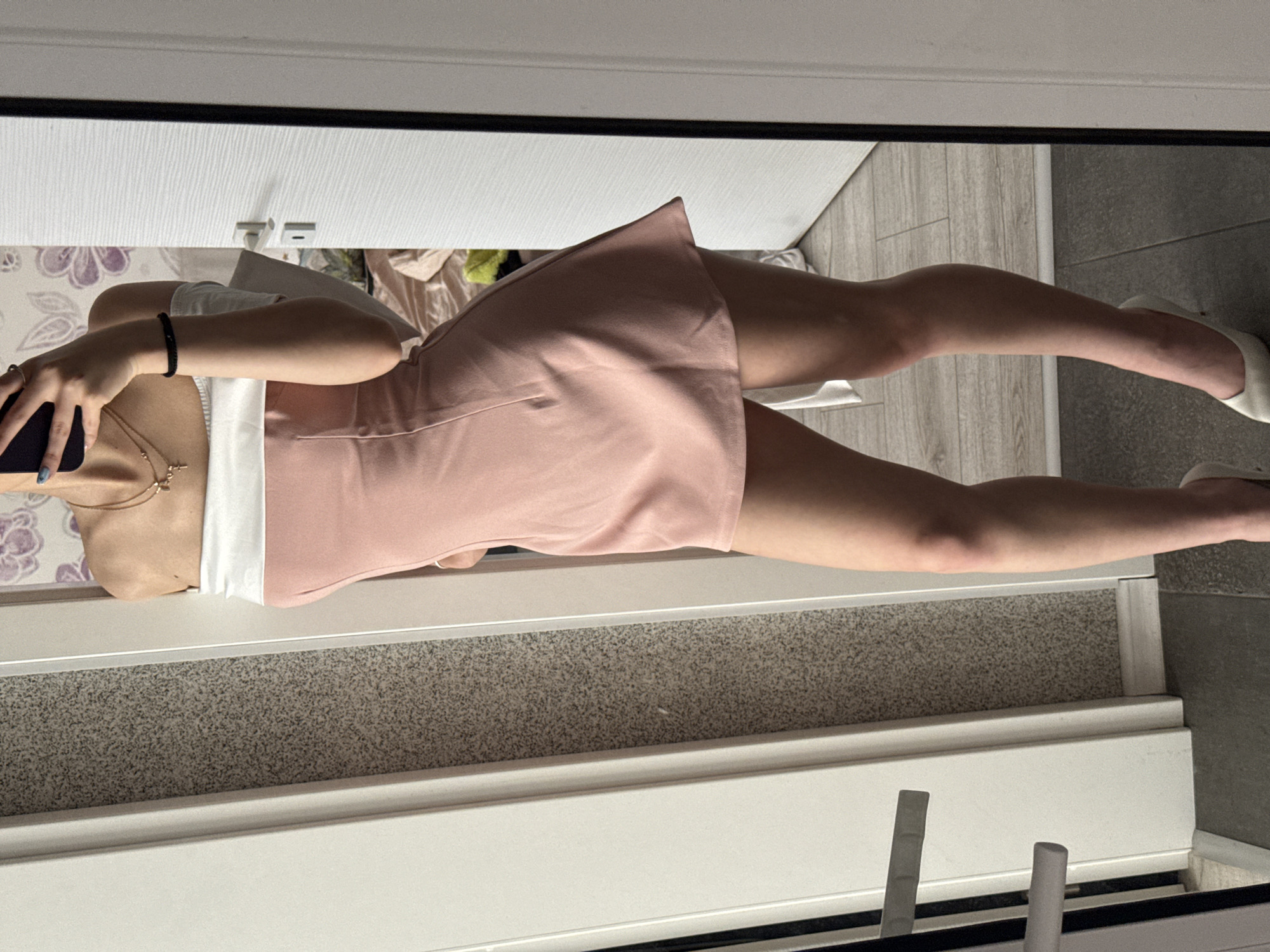 A medium shot presents a young woman in a peach skirt and white top, holding a smartphone, reflected in a mirror. The scene is indoors, with a gray and white carpeted area at the bottom of the frame. A cabinet or storage unit with visible door handles is at the top of the image, with hints of a floral pattern on what appears to be a bed or wall. A tiled floor suggests this might be a bathroom. Considering the style and interior, the location could potentially be within Sofia, Bulgaria, although specific landmarks are absent.