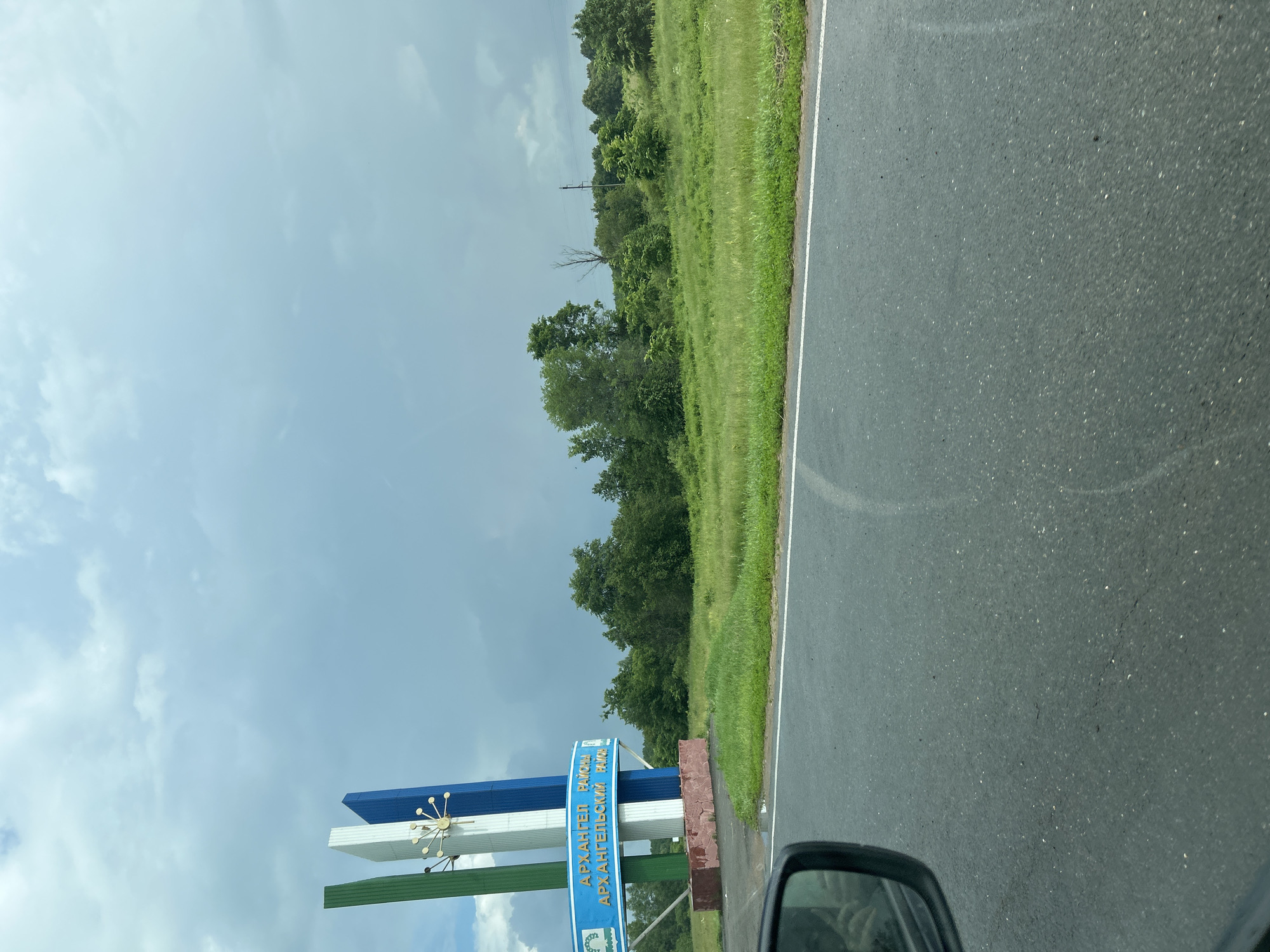 The image captures a roadside view in Irnykshe, Russia, featuring a prominent welcome sign. The sign is supported by three vertical columns, colored green, white, and blue, topped with a stylized, golden, starburst-like ornament. A blue banner across the lower part of the sign displays text in Russian, reading "АРХАНГЕЛ РАЙОНА" on the top line and "АРХАНГЕЛЬСКИМ РАЙОН" on the bottom line, indicating the Arkhangelsk region. The sign is anchored to a reddish-brown, stone-like base.
To the right of the sign, a verdant, grassy hill rises, densely populated with various trees. The sky above is overcast with heavy, grey clouds, suggesting an impending storm. In the foreground, a section of asphalt road is visible, with a white line marking the lane. The corner of a car's side mirror is visible in the lower left portion of the frame, reflecting a person's hand and arm. The overall impression is a rural landscape on a cloudy day, marking an entrance to the Arkhangelsk region.
