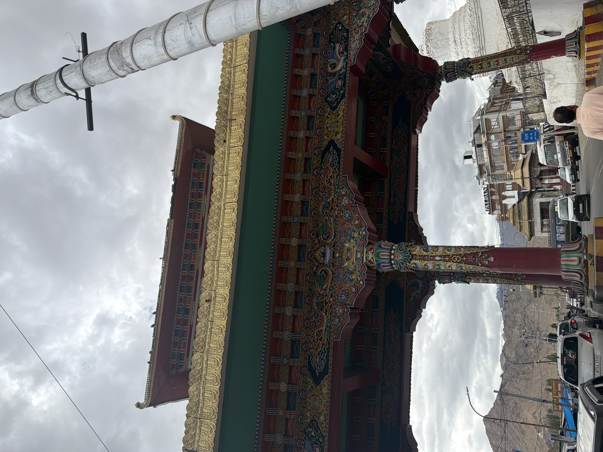 The image shows a wide, ornate gateway with traditional Tibetan architecture, painted in vibrant colors. The gateway is decorated with intricate patterns and designs, including mythical creatures and floral motifs. Gold trim is visible along the roofline and decorative elements. The gateway spans over a street filled with traffic, including cars and buses. In the background, there are buildings, including a multi-story structure with signs that appear to be for a hotel or business, and a white stupa. Mountains form the backdrop, with a cloudy, overcast sky overhead. The scene appears to be in a busy urban environment, likely a main thoroughfare in Leh, India, given the architectural style and mountainous landscape.