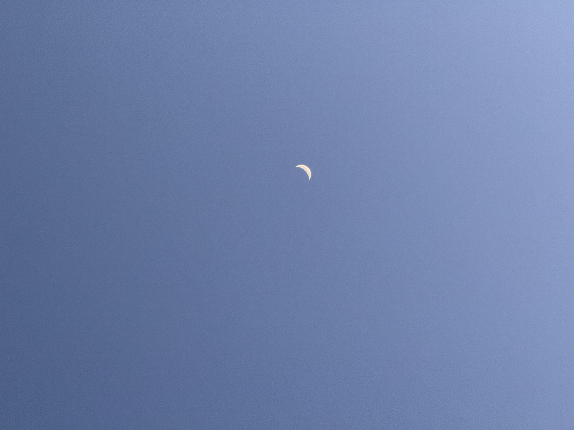 The image captures a clear, expansive blue sky, indicative of daytime conditions. A distinct, pale crescent moon is prominently featured in the upper central area of the frame. The sky is a uniform, vibrant blue, entirely devoid of clouds or any other atmospheric phenomena. The moon appears as a slender, luminous arc against the azure backdrop. There are no other celestial bodies, ground features, or man-made objects visible, making the sky and the moon the sole subjects. The scene conveys a sense of peacefulness and vastness. No specific activity or event is taking place beyond the static observation of the sky and moon. The time of day is clearly daytime, given the bright blue sky, with the moon being visible during daylight hours. The weather is clear and cloudless. No location cues are discernible within the image itself, though the provided context places this observation in Aksay, Kazakhstan. There is no visible text in the media file.