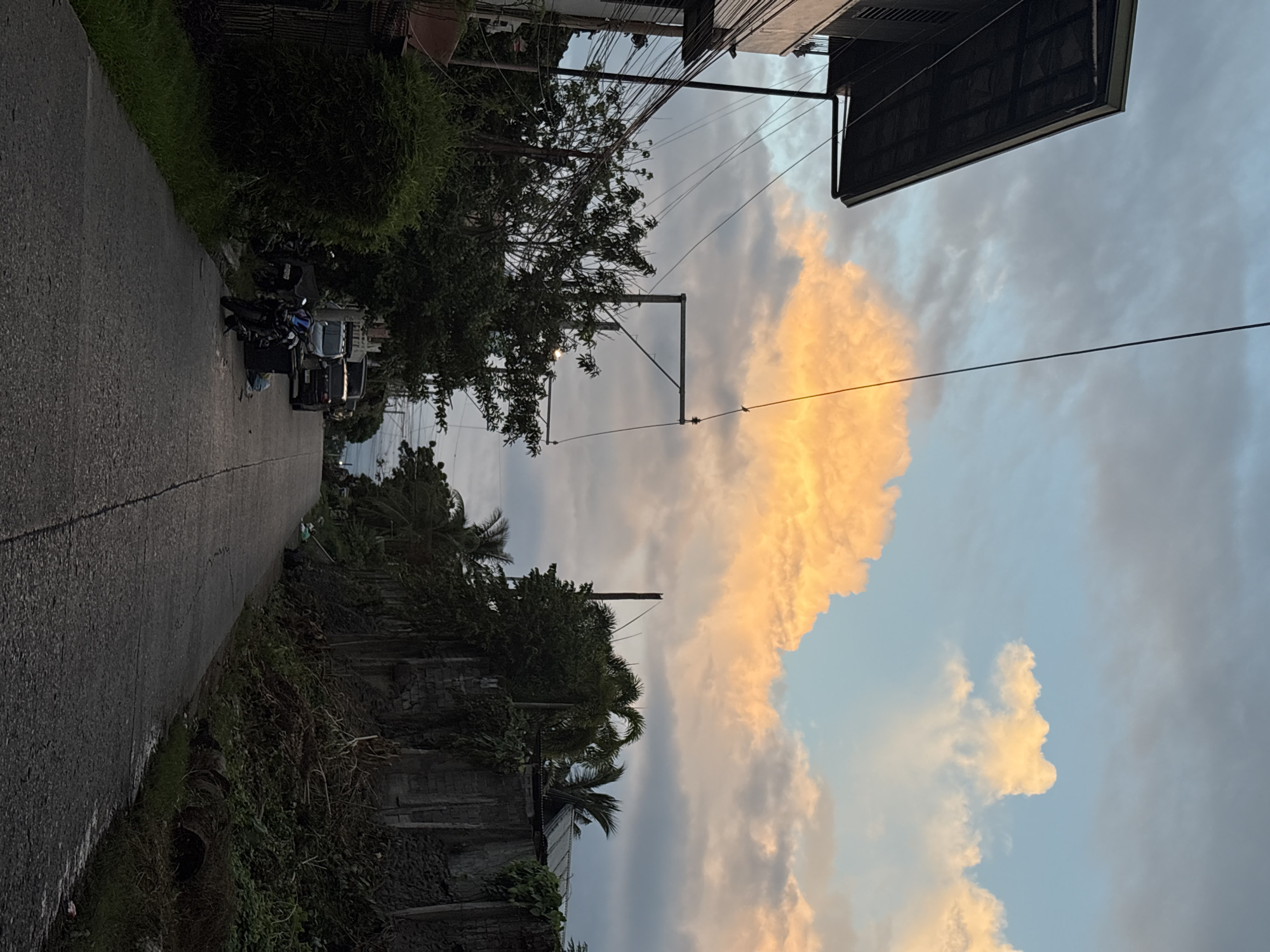 This is a medium-shot, eye-level, outdoor photograph taken in Davao City, Philippines, depicting a residential street during what appears to be dusk or dawn. The sky is a prominent feature, with a large, brightly lit orange cloud dominating the upper center, contrasting with a pale blue and grey sky with scattered, softer clouds. The street itself is paved with asphalt, leading into the distance, and appears to be slightly sloped.
On the left side of the street, a portion of a two-story building is visible, characterized by a dark, overhanging roof and a light-colored facade. Multiple electrical wires are strung across the scene, suspended from poles, adding to the urban feel. Adjacent to the building is dense green foliage, including bushes and trees. Several motorcycles are parked on the left side of the street, one of which is black with blue accents. Further down the street, a dark-colored SUV and a lighter-colored vehicle are parked.
On the right side of the street, a rough, stone-like wall runs along the pavement, partially covered in vegetation. Palm trees and other lush greenery are visible behind the wall, creating a natural barrier. The overall environment suggests a tropical setting, typical of Davao City. The lighting is soft, with the vibrant cloud casting an warm glow, indicating either sunrise or sunset. There are no people directly visible in the frame. The scene is quiet and still, conveying a sense of calm in a residential neighborhood.