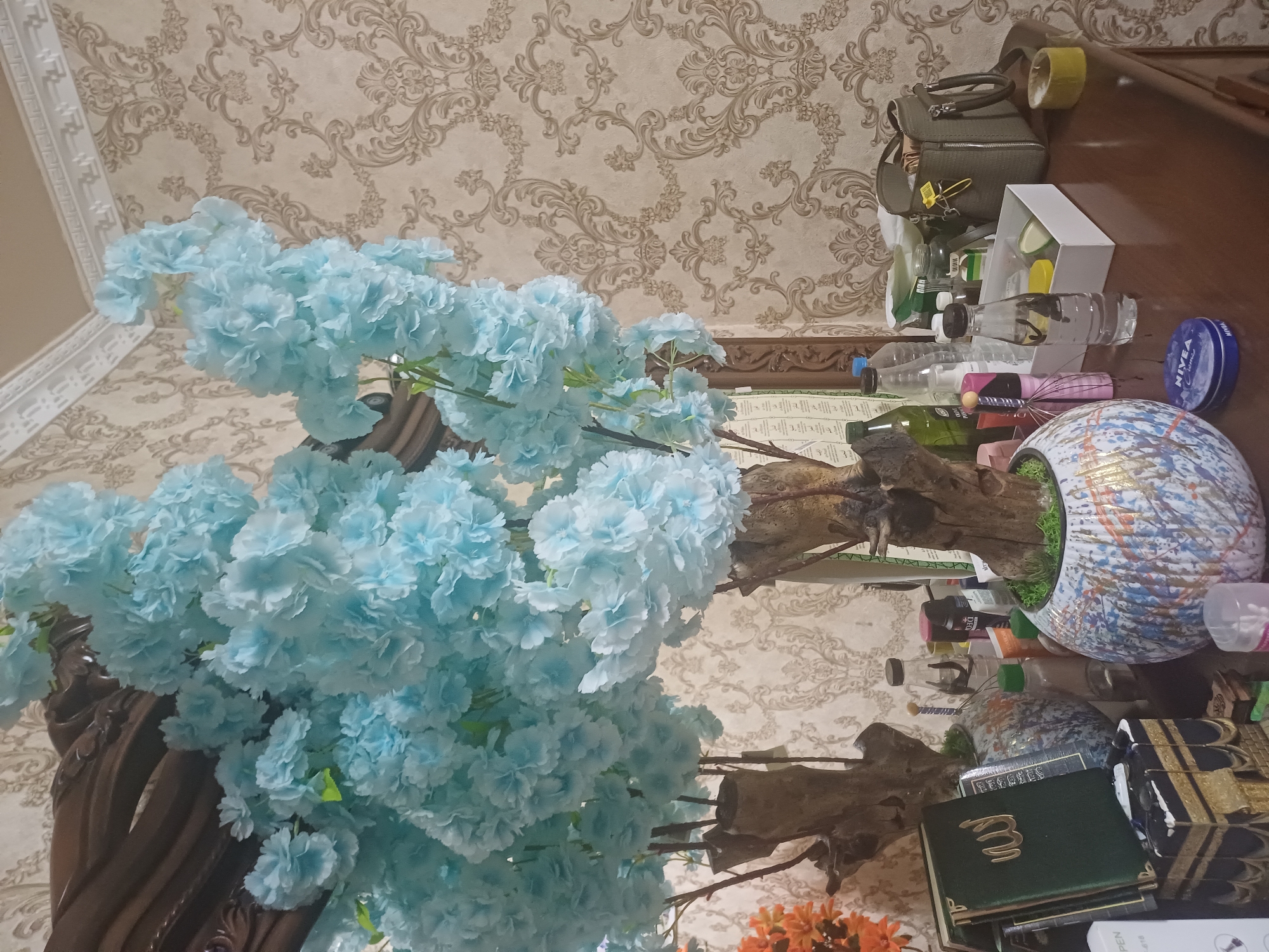 An indoor scene features a dark brown wooden dresser against a beige wallpaper with a damask pattern. Dominating the left side of the frame is a large, artificial tree with numerous light blue floral clusters. The tree's branches emerge from rough, exposed wooden bases set in decorative pots.
On the dresser, to the left, a dark green book with Arabic calligraphy is propped up. Next to it is a miniature replica of the Kaaba in Mecca. Scattered across the dresser are various items including bottles of liquids, small containers, a gray handbag, and a blue tin of Nivea cream. The lighting suggests an indoor setting, possibly a room in a residential or commercial space. There are no people visible in the image. The scene is likely staged for decorative purposes.
Given the information about the wallpaper and the general decor, this scene is consistent with an interior space in Tashkent, Uzbekistan, where such decorative elements and furnishings are common in homes and businesses.
