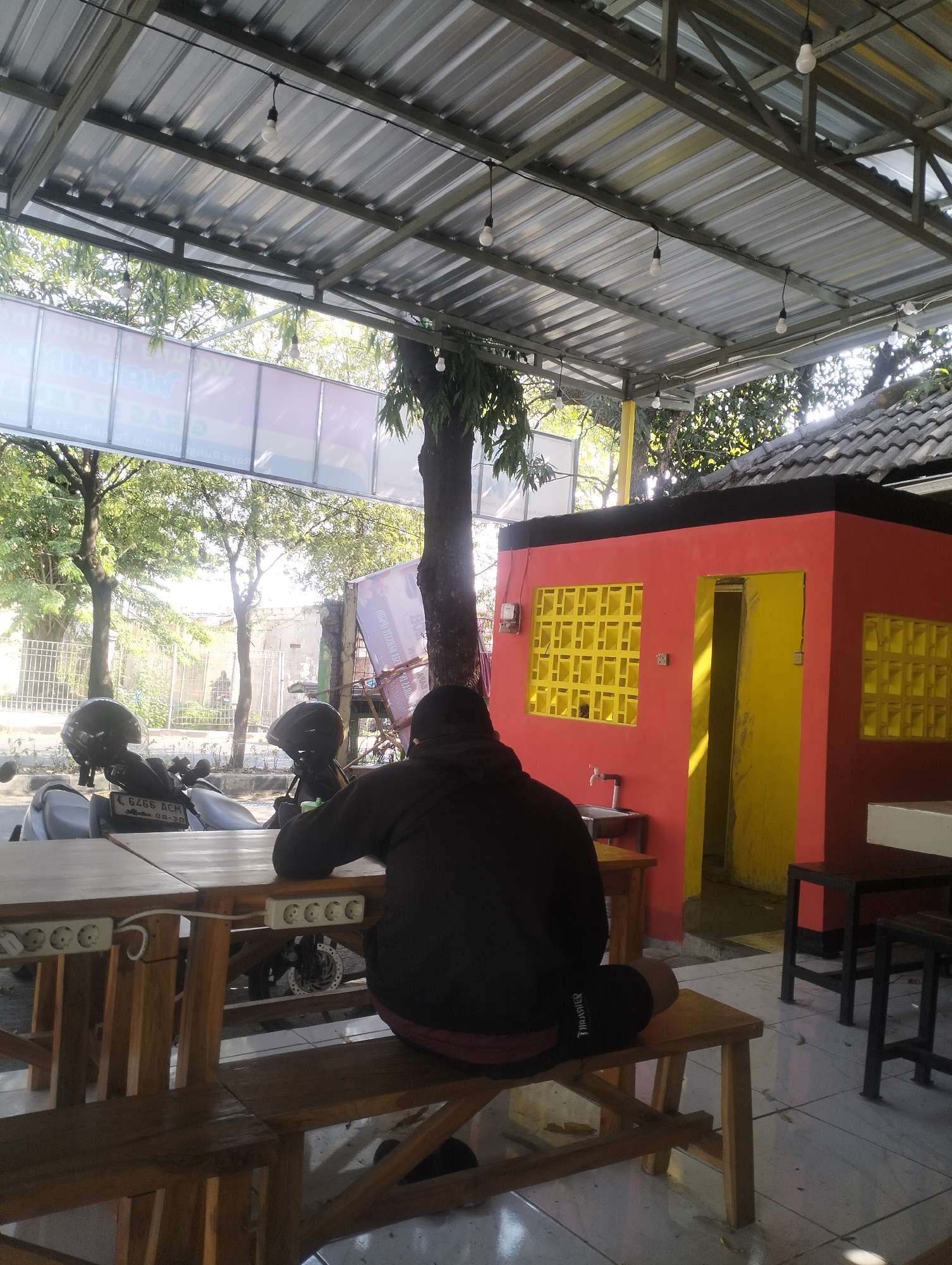 A person is sitting on a wooden bench at a table in an outdoor cafe. The person is wearing a black hooded jacket and black shorts. The cafe has a corrugated metal roof supported by metal beams. There is a red building with yellow windows and a yellow door in the background. Two motorcycles are parked behind the table. The cafe is located on a street in Surabaya, Indonesia.