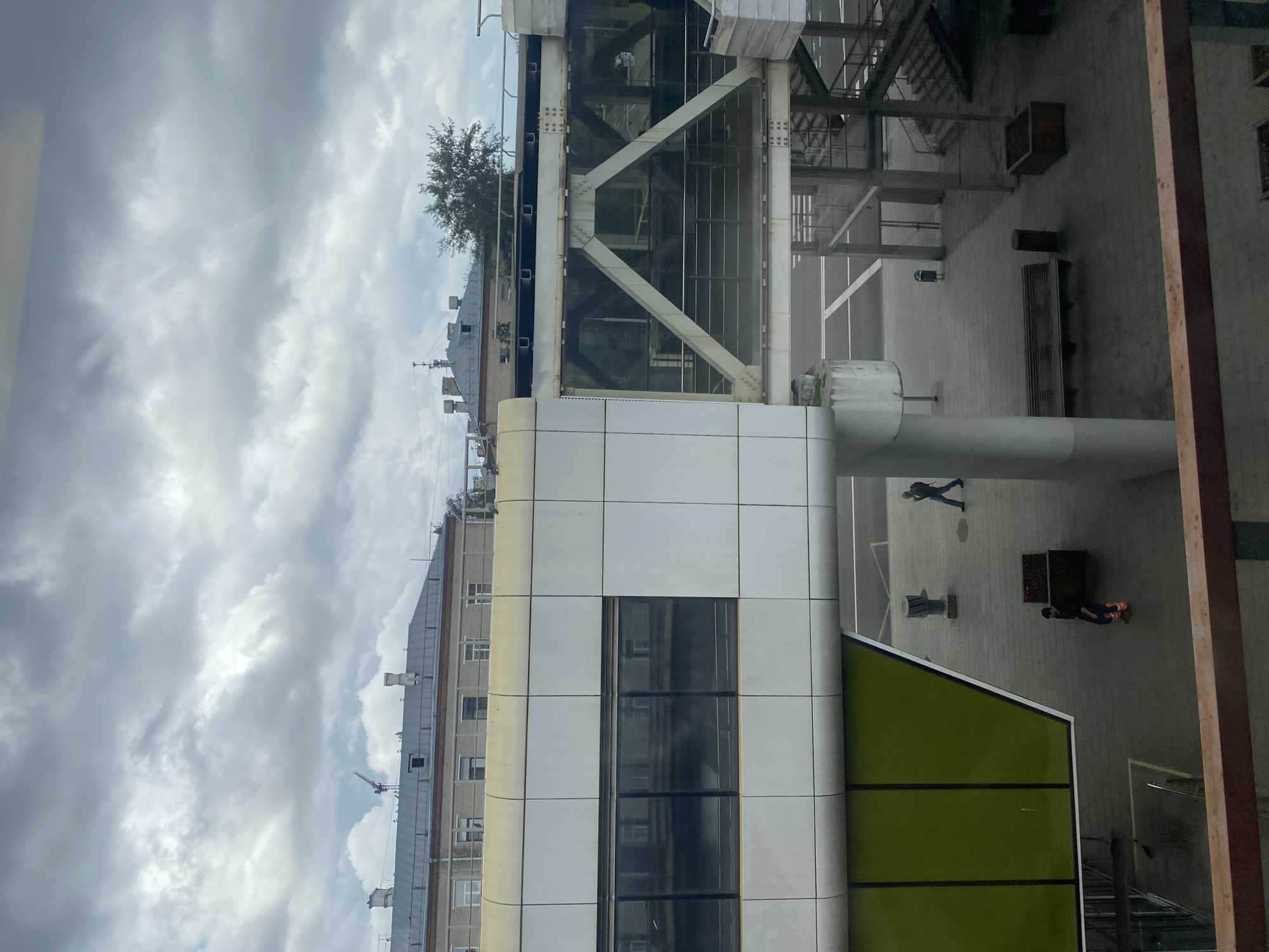 The image shows a modern building with a glass skywalk. The skywalk is supported by a white triangular structure and connects to another building. In the foreground, there is a green wall and a brown railing. Outside the building, there is a paved area with benches, trash cans, and a staircase. Two people are visible walking in the paved area. The sky is cloudy, and the overall atmosphere is overcast. The image appears to have been taken from a window. This location is likely the Moscow Metro station or an airport terminal.