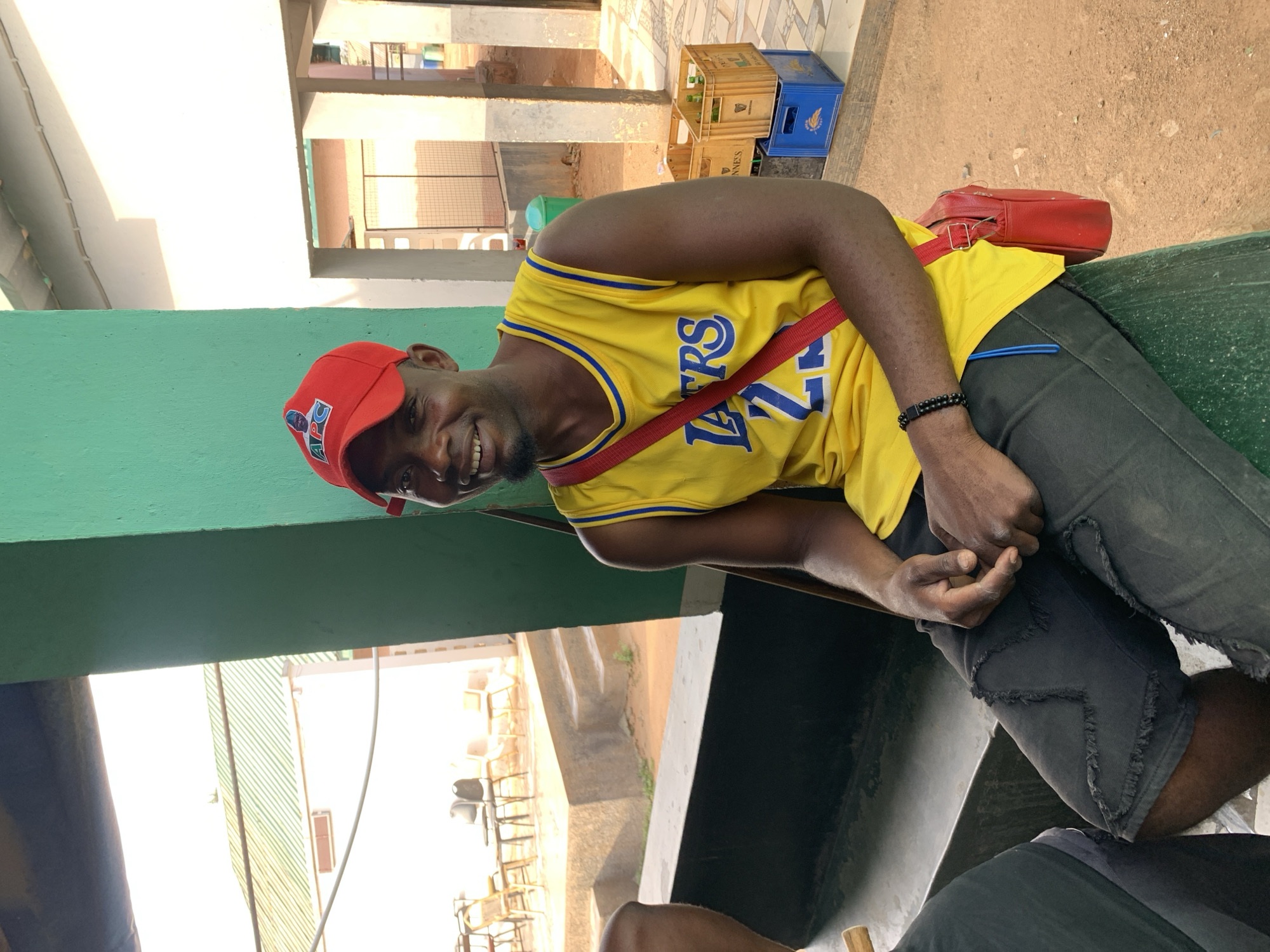 A man is sitting outdoors, wearing a yellow Lakers jersey with blue trim and the number 24, a red baseball cap with an emblem on it, and black denim shorts. He has a red strap across his chest, supporting a red bag. He is seated on a green surface. Behind him are partially visible buildings with neutral color tones. In the distance, wooden crates and a corrugated metal structure can be seen. The image was captured during daylight hours. The subject is smiling. The location is New Stadium, Nigeria.