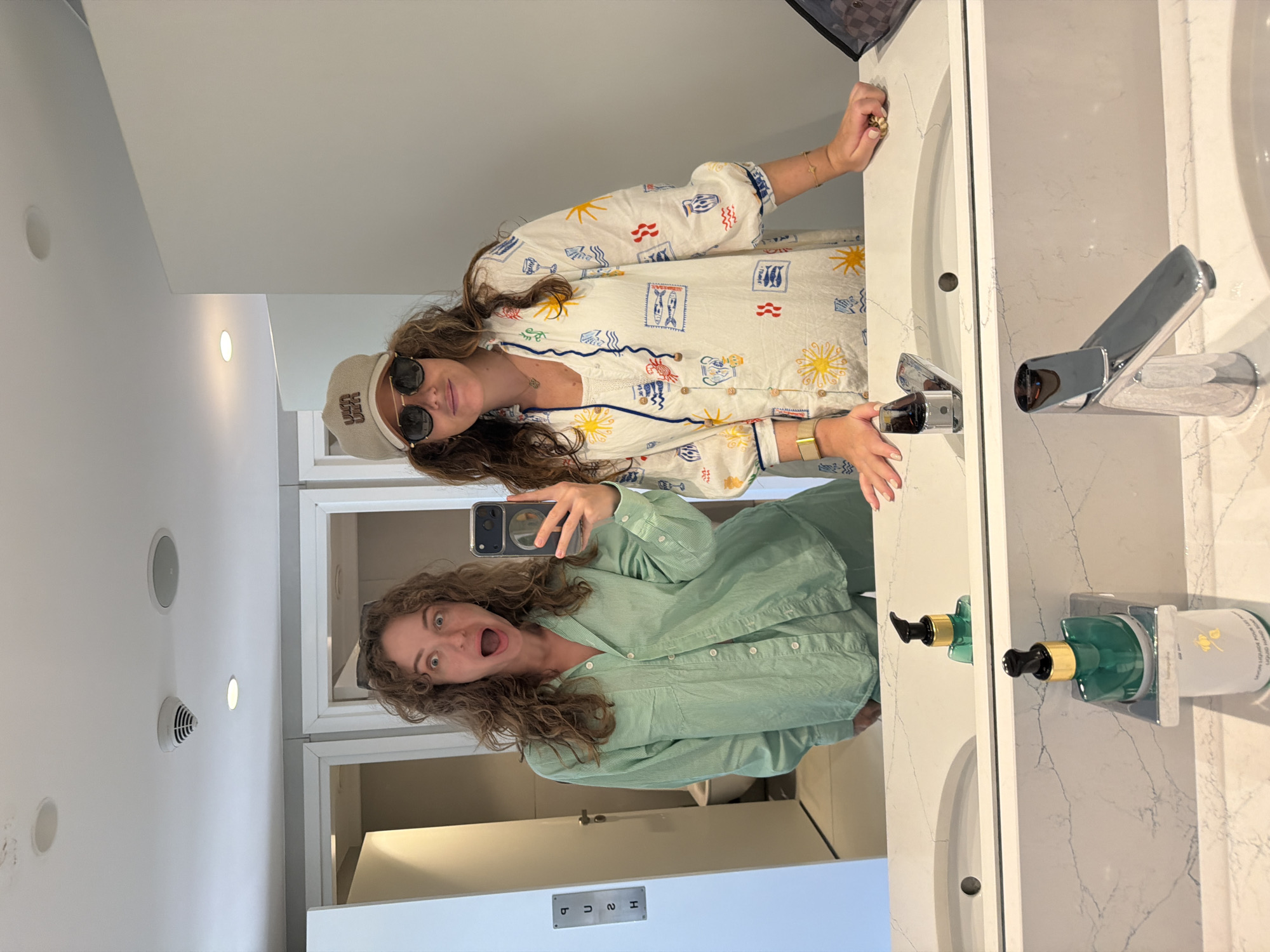 Two young women are taking a mirror selfie in a modern bathroom with white marble countertops. The woman on the left is wearing a mint green pajama set and has a surprised expression with her mouth open. She is holding a smartphone in her right hand, which is angled to capture the reflection. The woman on the right is wearing a white pajama top with a colorful print of suns, fish, and other nautical motifs. She has on sunglasses and a tan baseball cap. Her left arm is resting on the countertop, and her right hand is visible, showing a bracelet and rings. The bathroom features two sinks, chrome faucets, and soap dispensers. A "PUSH" sign is visible on a door in the background. The overall impression is that of a casual, personal moment shared between friends in a well-appointed private space.