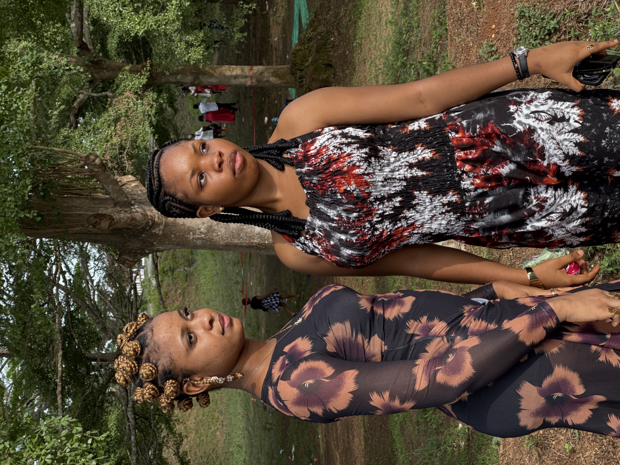 Two women stand in an outdoor setting in Ihiagwa, Nigeria. The woman closer to the camera wears a long-sleeved, sheer black top with large brown flower patterns, her hair styled in small buns. She also wears gold jewelry: a necklace, earrings, rings, and a bracelet. The second woman wears a sleeveless dress with a black, white, and red pattern and braided hair. She wears a watch and bracelets on her left wrist. A pair of glasses and a black phone or wallet are in her left hand. The background includes trees, grass, and other people.