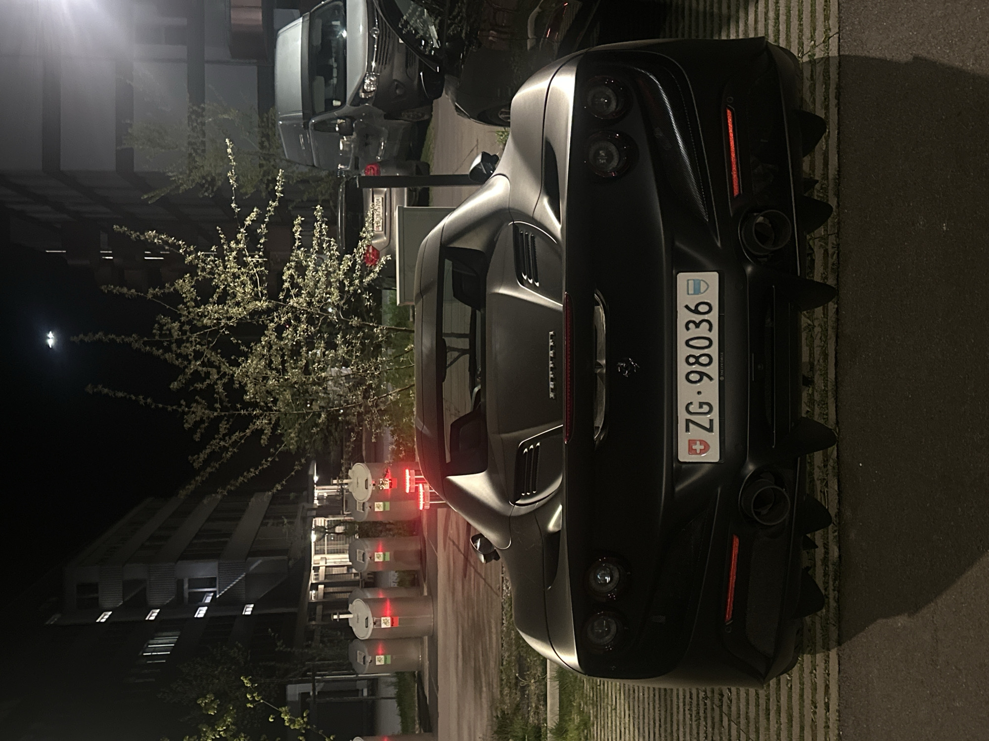 The image presents a dark, nighttime scene in Dübendorf, Switzerland, featuring a black luxury sports car, specifically a Ferrari, parked outdoors. The car is viewed from the rear, highlighting its sleek body, four circular taillights, and a prominent rear diffuser with quad exhaust pipes. A Swiss license plate, "ZG-98036," is centrally affixed to the car's rear, with the small text "THE LIVE CIRCLE" visible on its bottom edge. The "FERRARI" brand name is also visible on the trunk lid.
In the background, other parked vehicles are partially visible, including a light-colored SUV or van and a darker car. The scene is illuminated by artificial light sources, creating reflections on the cars and casting a glow on a nearby flowering tree or bush adorned with numerous white blossoms. Further in the background, dimly lit buildings with some illuminated windows are discernible against the dark night sky. The ground appears to be a paved surface, likely a parking area, with subtle striped markings. Several cylindrical, light-colored objects, possibly waste containers or urban furniture, are lined up to the left of the scene. The image captures a static, urban nighttime environment with the high-performance vehicle as its central subject.