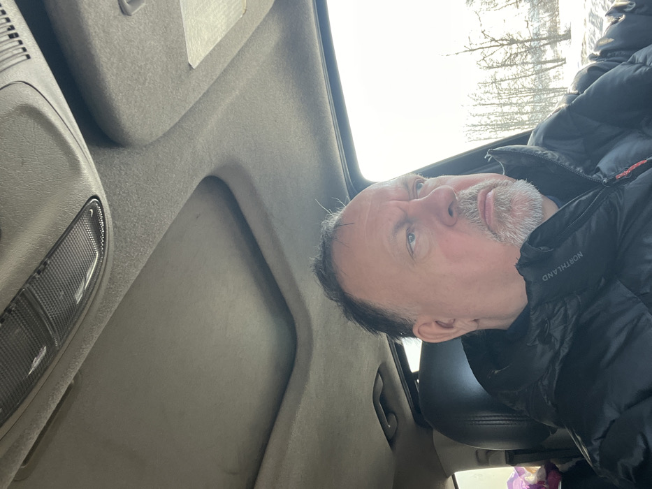 The image captures a middle-aged male seated inside a vehicle, facing and looking towards the right side of the frame. He has dark hair and a grey goatee, wearing a black puffer jacket with "NORTHLAND" visible on the left chest. The vehicle's interior features a light-colored headliner, an overhead console with a rectangular light fixture, and a visible sunroof opening. Through the vehicle's window, a bright, snowy landscape is visible, showing snow-covered ground and bare deciduous trees or shrubs under a bright sky, indicative of a winter season. The scene takes place in Ерино, Russia.