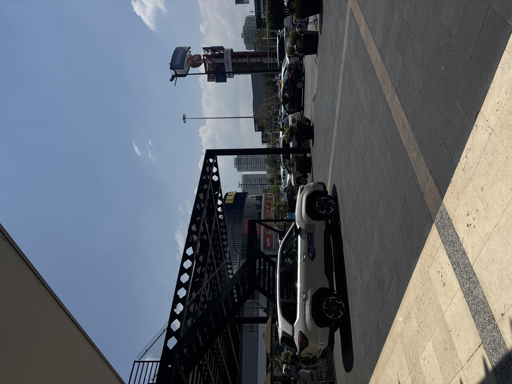 The image shows an outdoor scene, possibly in San Bernardino Tlaxcalancingo, Mexico, as suggested by the "Via San Angel" sign on a tower that also features a "Walmart" sign. The image appears to be taken on a sunny day with a blue sky and some clouds.
In the foreground, there is a paved area with different tile patterns. A white Subaru SUV is parked. Part of a dark metal structure with hexagonal cutouts is visible to the left of the vehicle. Further back, several parked cars can be seen in a row.
In the background, there are buildings and skyscrapers. One building has what appears to be a portion of an IKEA logo on it. Streetlights are also visible. People can be seen in the background near the base of the metal structure.