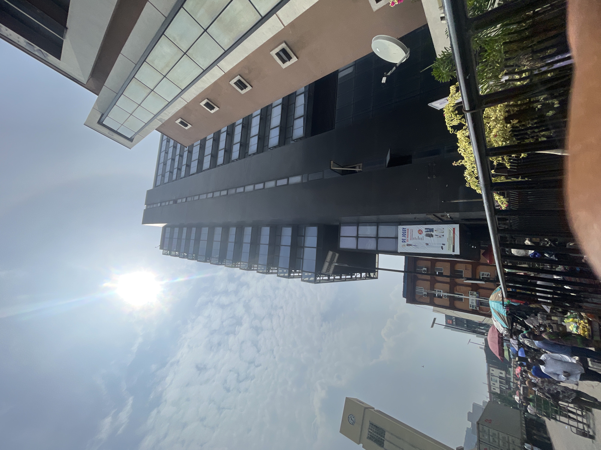 The image shows a street scene in Lagos, Nigeria, with a focus on the architecture and street activity. The camera is angled upwards, capturing a view of tall buildings with glass windows and a mix of dark and light facades. The sky is bright, with a visible sun and scattered clouds.
On the right side of the frame, there is a bustling street scene with people walking and interacting, suggesting a crowded market or commercial area. There are visible umbrellas and makeshift stalls, indicating outdoor vendors.
A clock tower is also visible lower center.