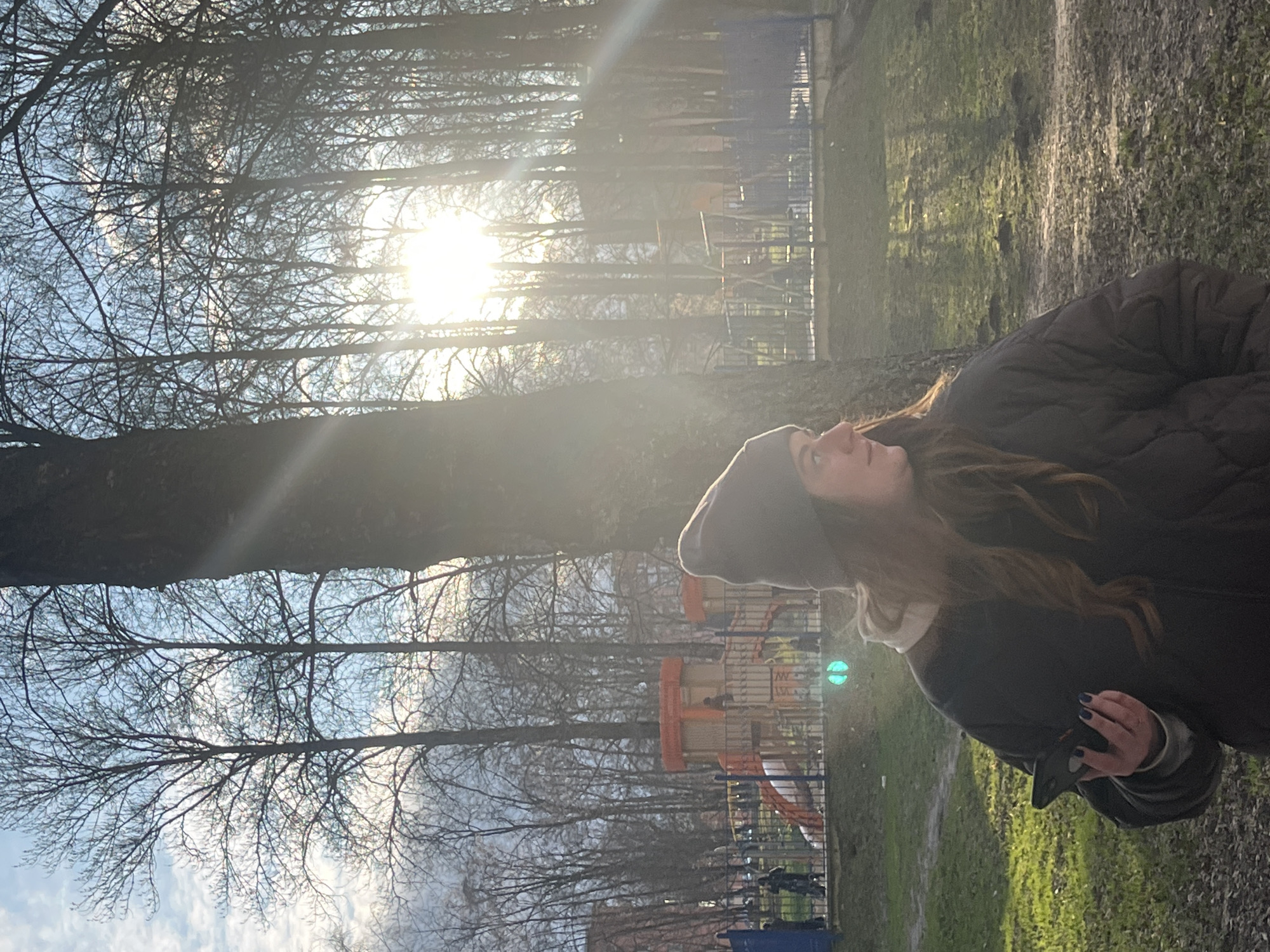 The image shows a woman in an outdoor setting, likely a park in Klin, Russia. She is wearing a dark beanie and a puffy dark jacket. She is holding a dark object in her hand. The background features trees without leaves and the sun shining through the branches. A playground structure can be seen in the distance, as well as a patch of green grass. The environment appears to be a public park in the city of Klin, Russia.