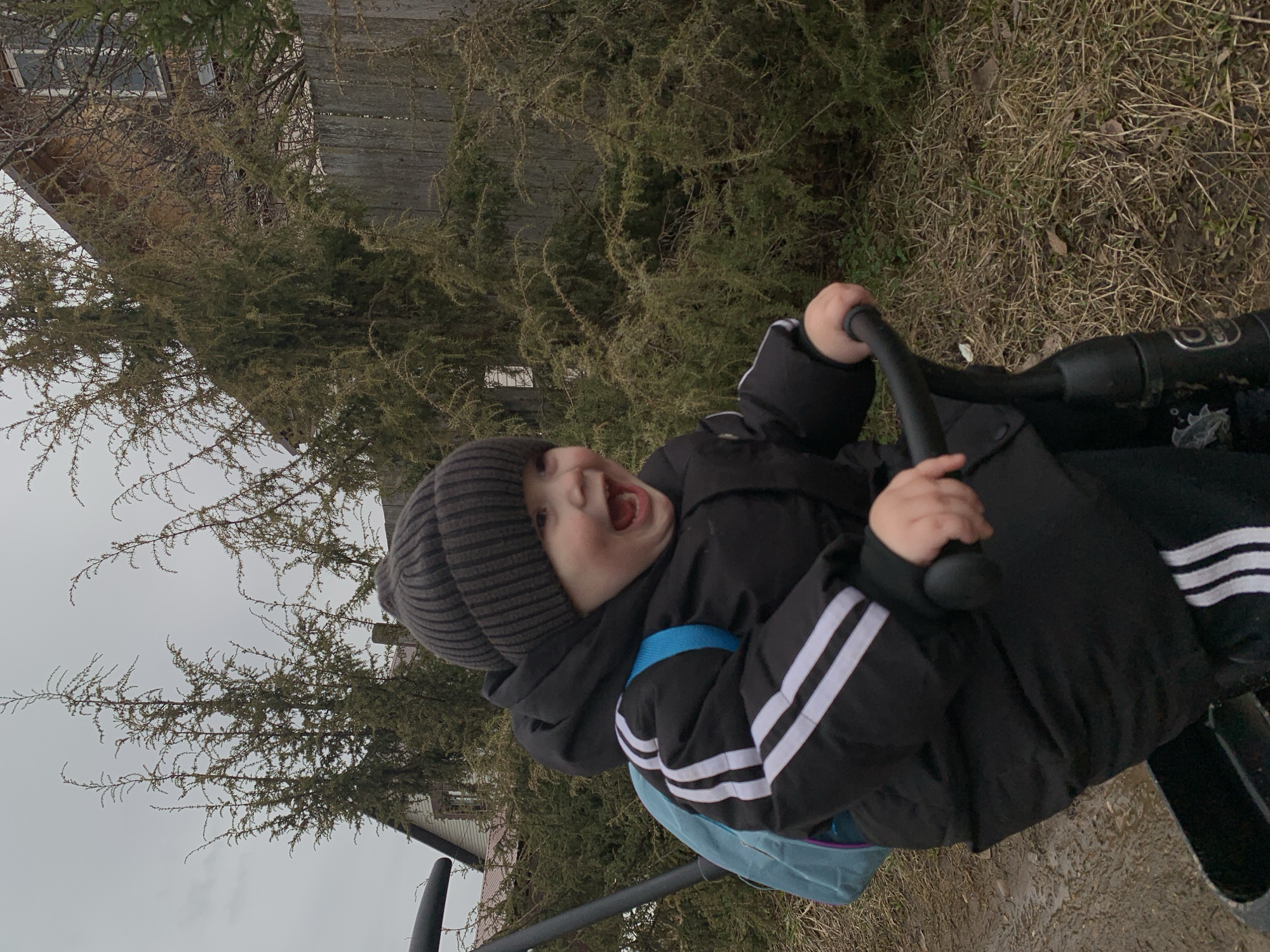 A young child wearing a gray striped beanie, a black jacket with white stripes on the sleeves, and a light blue backpack is on a swing. The child has an open mouth as if laughing or shouting. The background includes dry grass, coniferous bushes or trees, and a wooden building. Based on the location Фоки, Russia, the environment suggests a suburban or rural residential area.