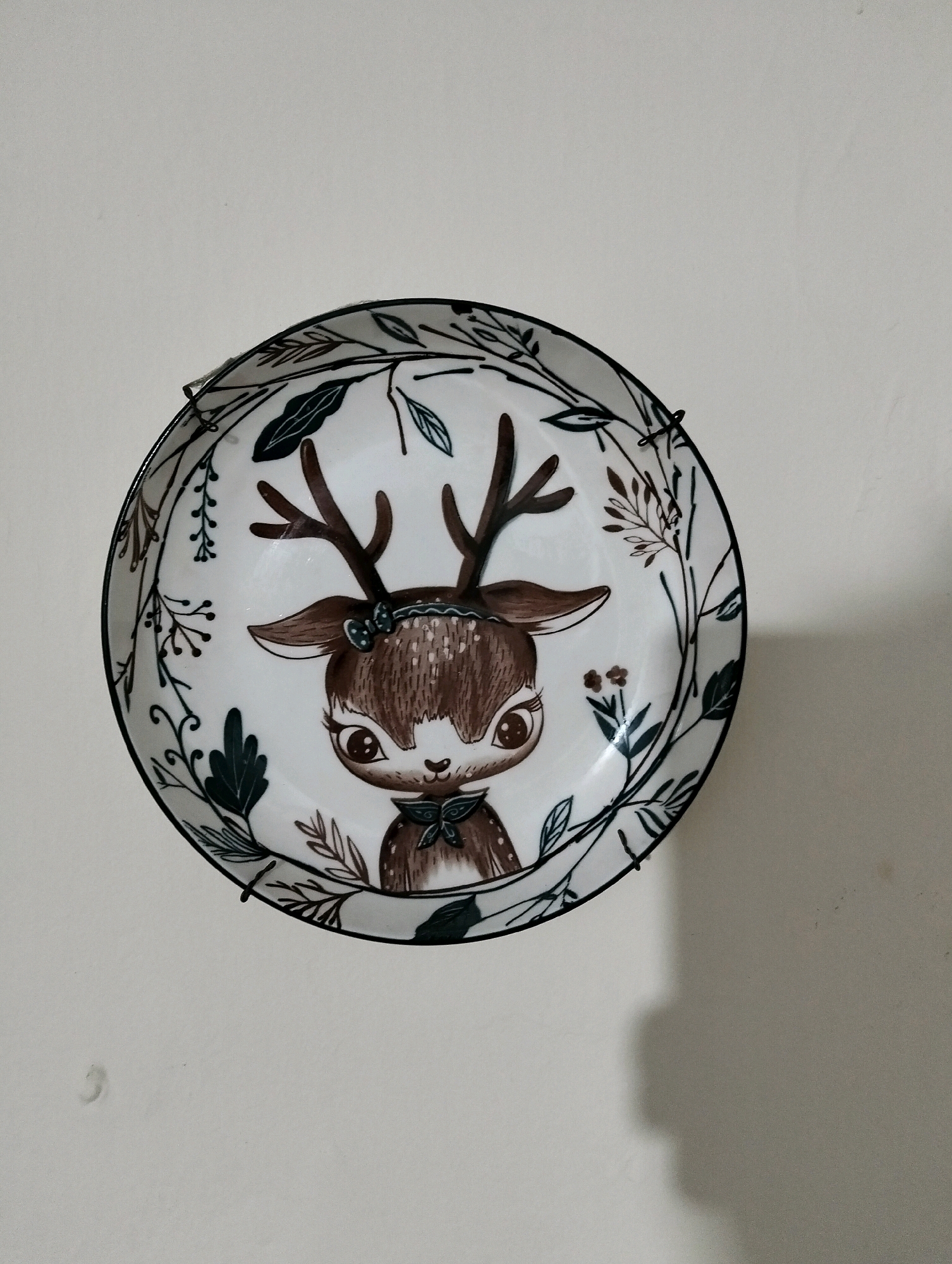 A decorative ceramic plate with a cartoon deer illustration is mounted on a light-colored wall in Patrol, Indonesia. The plate features a white background with a border of stylized green and brown leaves and branches. In the center, a young deer with large, expressive eyes and small antlers is depicted. The deer is wearing a green bow around its neck and a blue bow on its head. The plate is held in place by black wire plate hangers. A shadow of the plate is cast on the wall to the right.