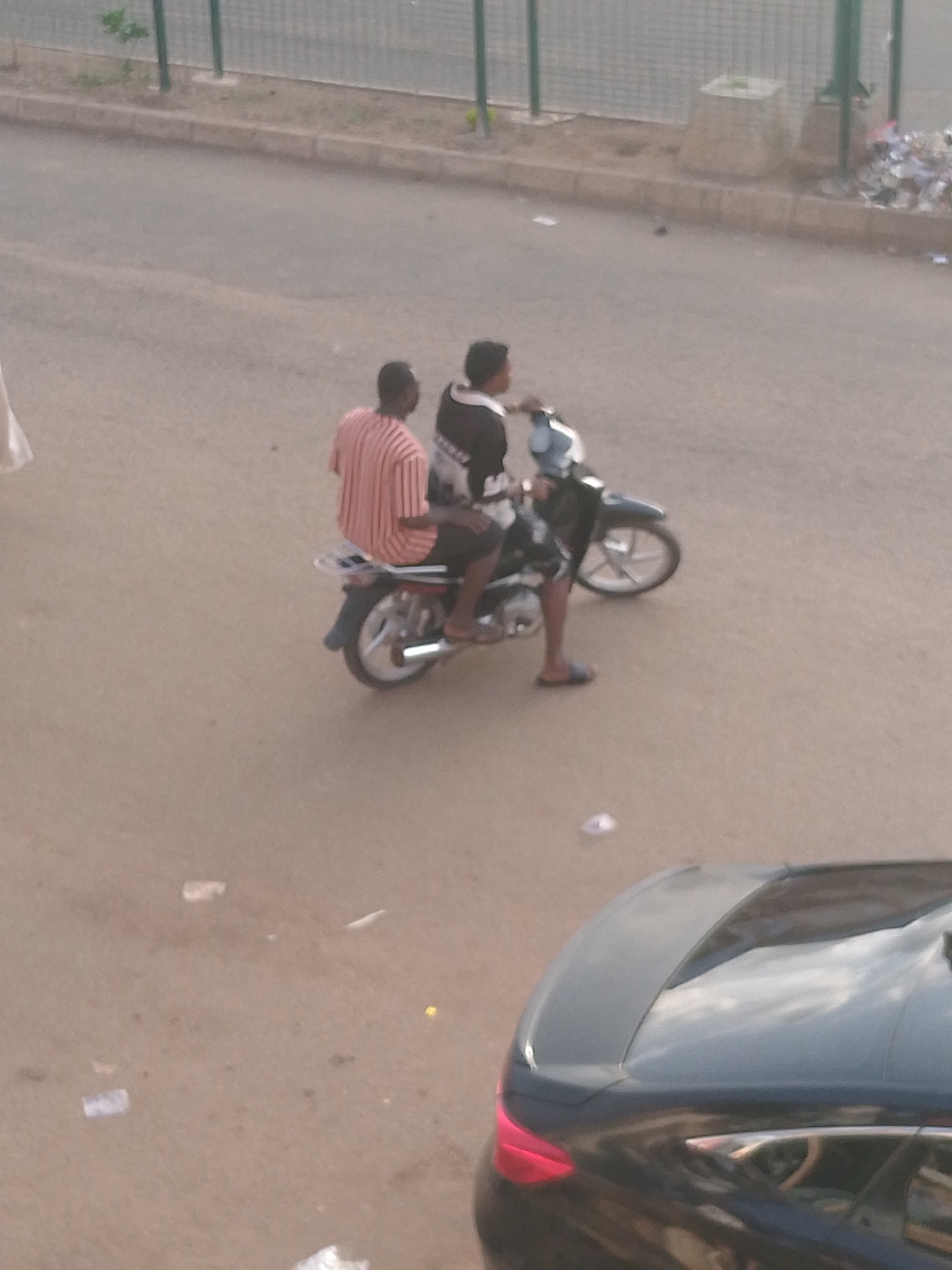 In Bukuru, Nigeria, two individuals are riding a motorcycle on a paved road. The person in the front is wearing a striped short-sleeved shirt, and the person behind them is wearing a black and white patterned shirt. They are both facing away from the viewer. The motorcycle is dark colored with a silver rack on the back. A dark-colored car is visible in the foreground, partially obscuring the lower portion of the image. The road appears to be somewhat dirty with scattered debris. In the background, a metal fence borders the road, with a trash can and a pile of refuse visible beyond it. The scene is lit by daylight.