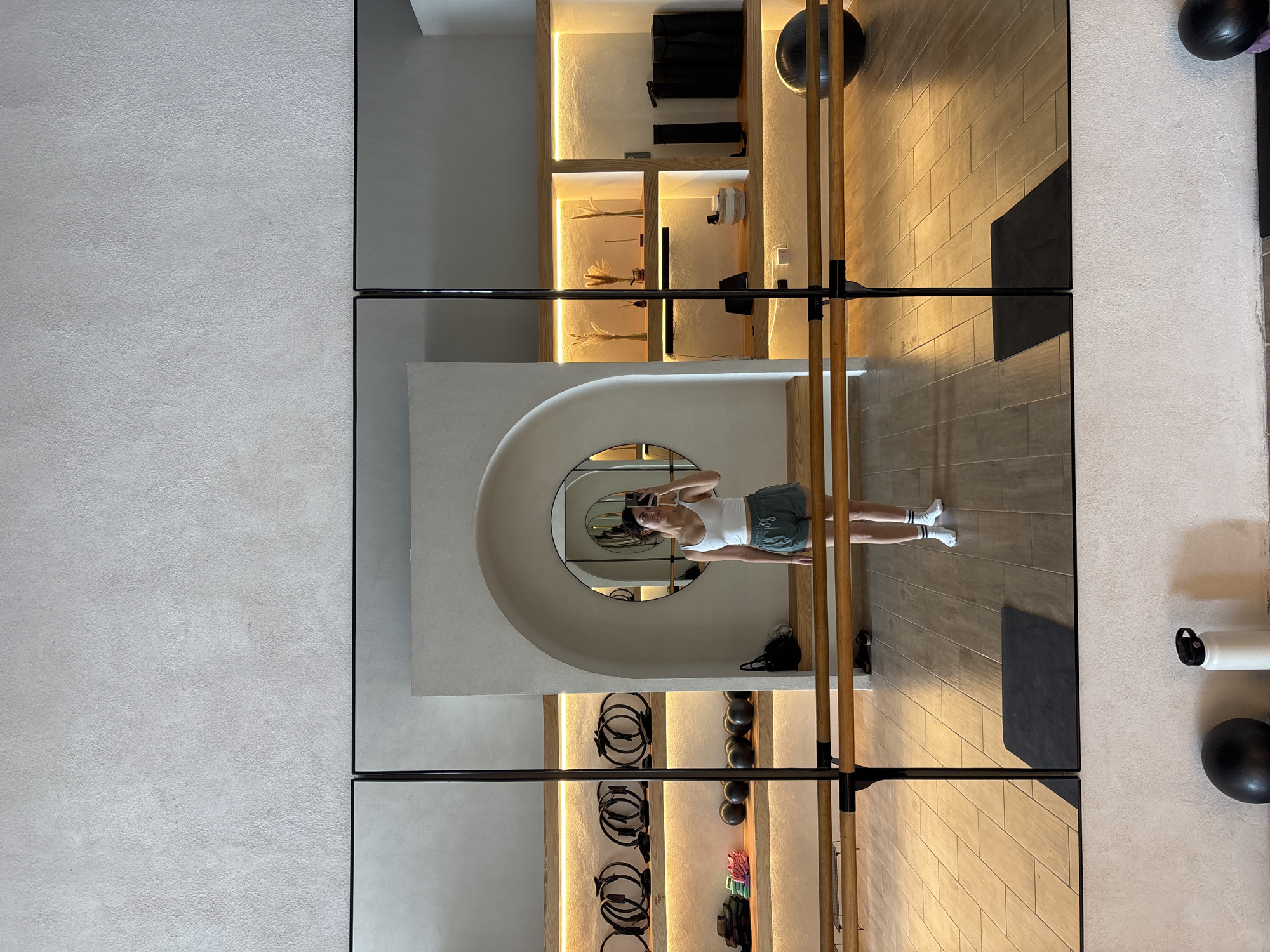 The image depicts an interior scene, likely in a fitness studio or gym in San Bernardino Tlaxcalancingo, Mexico, judging by the presence of equipment like pilates rings, fitness balls, and a ballet barre. The scene is captured through a series of large mirrors, reflecting the interior space and a woman taking a selfie. The woman is wearing a tank top and shorts, with white socks. The room has built-in shelving with indirect lighting, and the floors appear to be tiled. The overall aesthetic suggests a modern and well-maintained facility.
