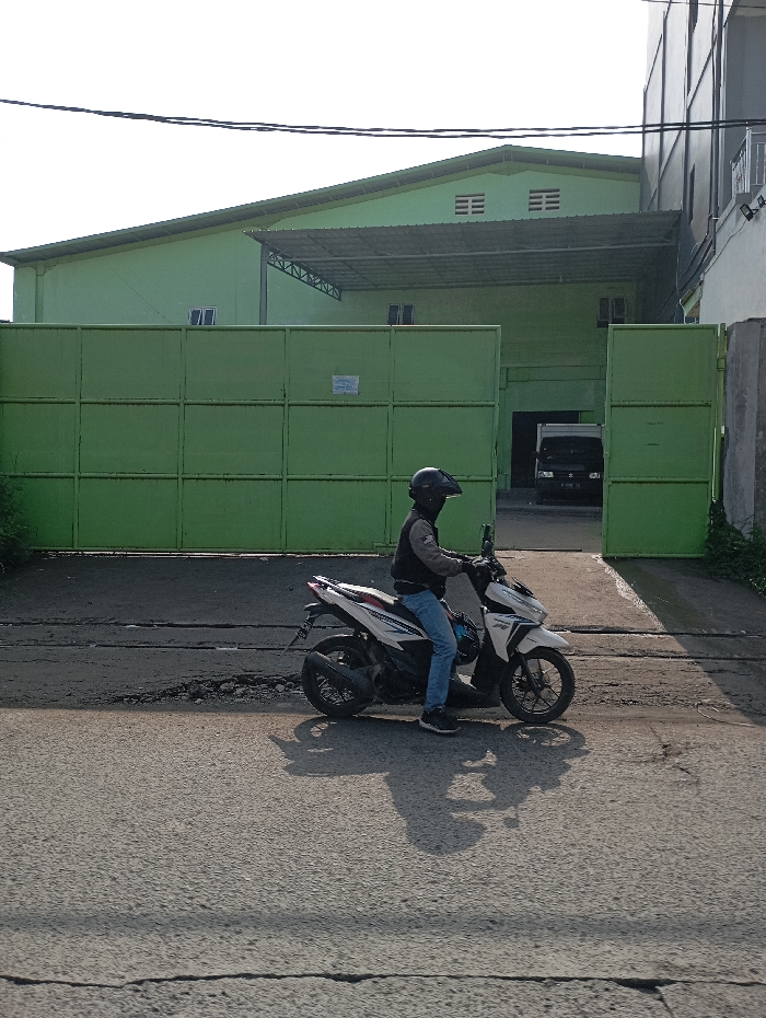 An individual sits astride a white and black scooter-style motorcycle on an asphalt road in Pataruman, Indonesia. The person wears a dark full-face helmet with a clear visor, a dark jacket with grey sleeves, blue jeans, and dark athletic shoes, with both feet resting on the ground. A prominent shadow of the person and motorcycle extends to the left, indicating direct sunlight from the right. In the background, a large, light green industrial building with a metal roof and an extensive matching green panel gate is visible. The gate is partially open on the right, revealing a dark-colored truck parked within the facility. Portions of another building are visible to the far right. Overhead power lines traverse the top of the scene against a clear sky. The road surface exhibits signs of wear and cracking.