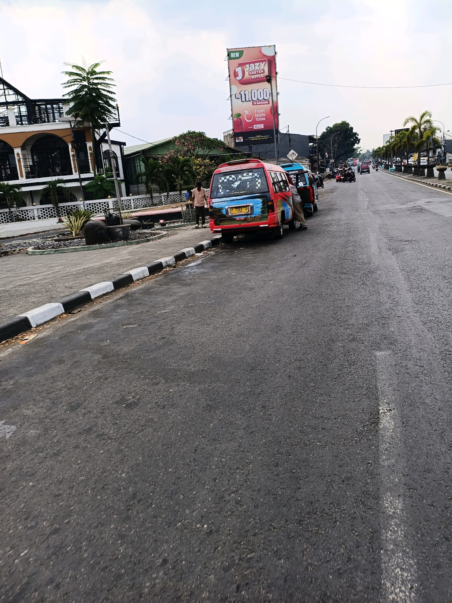 The image depicts a street scene in Sukabumi, Indonesia, with a focus on a row of parked vehicles and buildings lining the road. In the foreground, a black and white striped curb runs along the edge of the asphalt road, which appears to be wet in some areas, suggesting recent rain.
Dominating the left side of the frame is a multi-story building with a distinctive arched facade on the lower level and a glass-enclosed upper story. Adjacent to this building is a vibrant red and blue mini-bus, its rear facing the camera, displaying a license plate. Another mini-bus, blue in color, is parked next to it. A person in a light-colored shirt and pants stands next to the red and blue mini-bus, appearing to interact with it. Further down the road, more vehicles, including motorbikes and other mini-buses, are visible, some in motion.
To the right of the mini-buses, a large billboard stands tall, advertising "Jazy Kretek Apple" with a price of 11,000. Palm trees are planted along the side of the road, adding to the tropical setting. The sky above is overcast with clouds. The overall impression is that of a busy street in a Southeast Asian city, likely during the day.