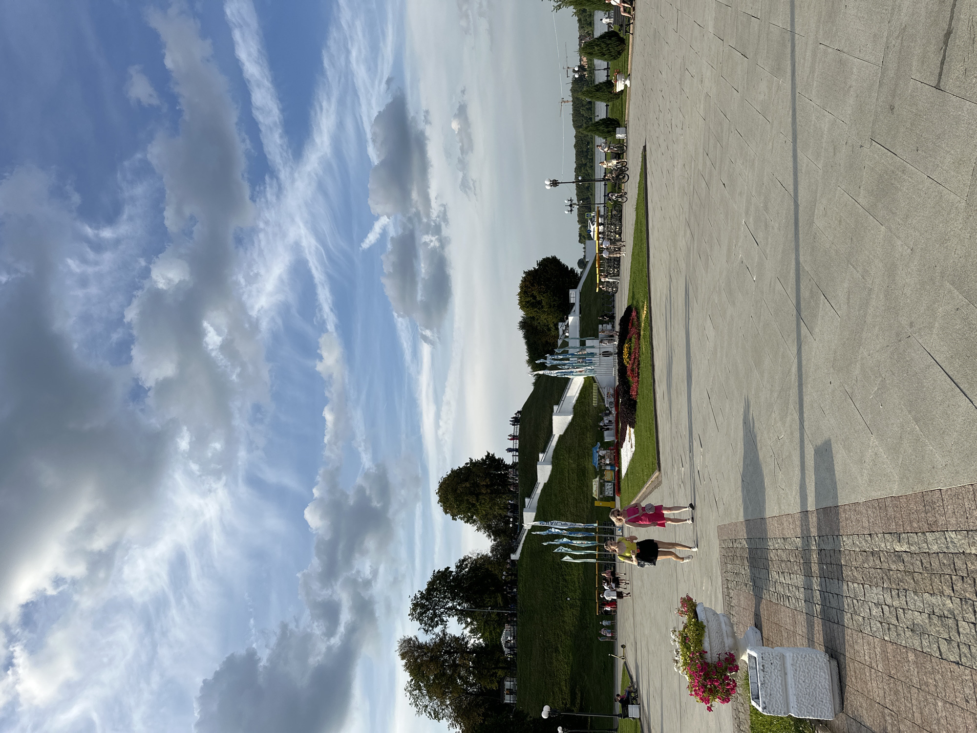 The image depicts a daytime scene at a park or promenade in Rusanovo, Russia. The sky is a prominent feature, filled with dramatic clouds and streaks of lighter clouds, suggesting a dynamic weather pattern. A paved walkway occupies the foreground, with people walking and sitting along its edges.
Further into the scene, a lush green lawn slopes upwards, dotted with trees. Along the slope, there are decorative elements, including a series of flags or banners displayed on poles. A white, modern-looking structure with multiple tiers is also visible on the grassy incline. At the bottom of the slope, there's a vibrant flowerbed with red and yellow blooms.
Two young women are the most prominent subjects in the foreground, walking on the paved area. One is wearing a pink dress, and the other a yellow top and black skirt. Their posture and proximity suggest they are together. In the background, several other individuals and groups are scattered throughout the park, some appearing to be cycling or sitting. The presence of bicycles suggests a recreational area.
The lighting indicates it is likely late afternoon or early evening, given the long shadows cast by the people and objects. The overall atmosphere appears relaxed and pleasant, consistent with a public park on a day with interesting cloud formations. No specific text is clearly legible in the image.