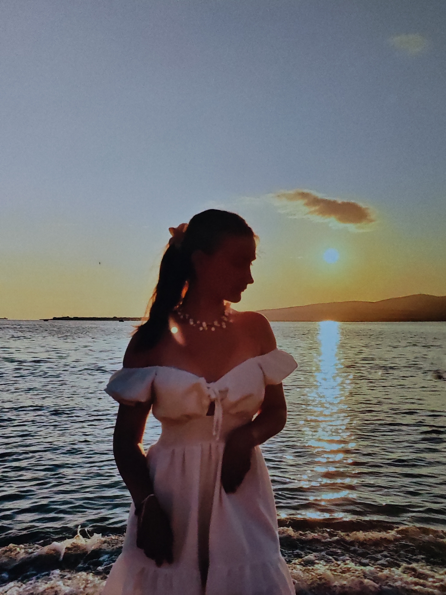 A silhouette of a young woman stands on the edge of a body of water during sunset. She is wearing a white off-the-shoulder dress with puff sleeves and a pearl necklace. Her hair is pulled back in a ponytail with a clip. The sun is setting over a distant mountain range, casting a golden light on the water and the sky. There are small waves lapping at the shore where she is standing. The overall scene evokes a sense of peace and tranquility.