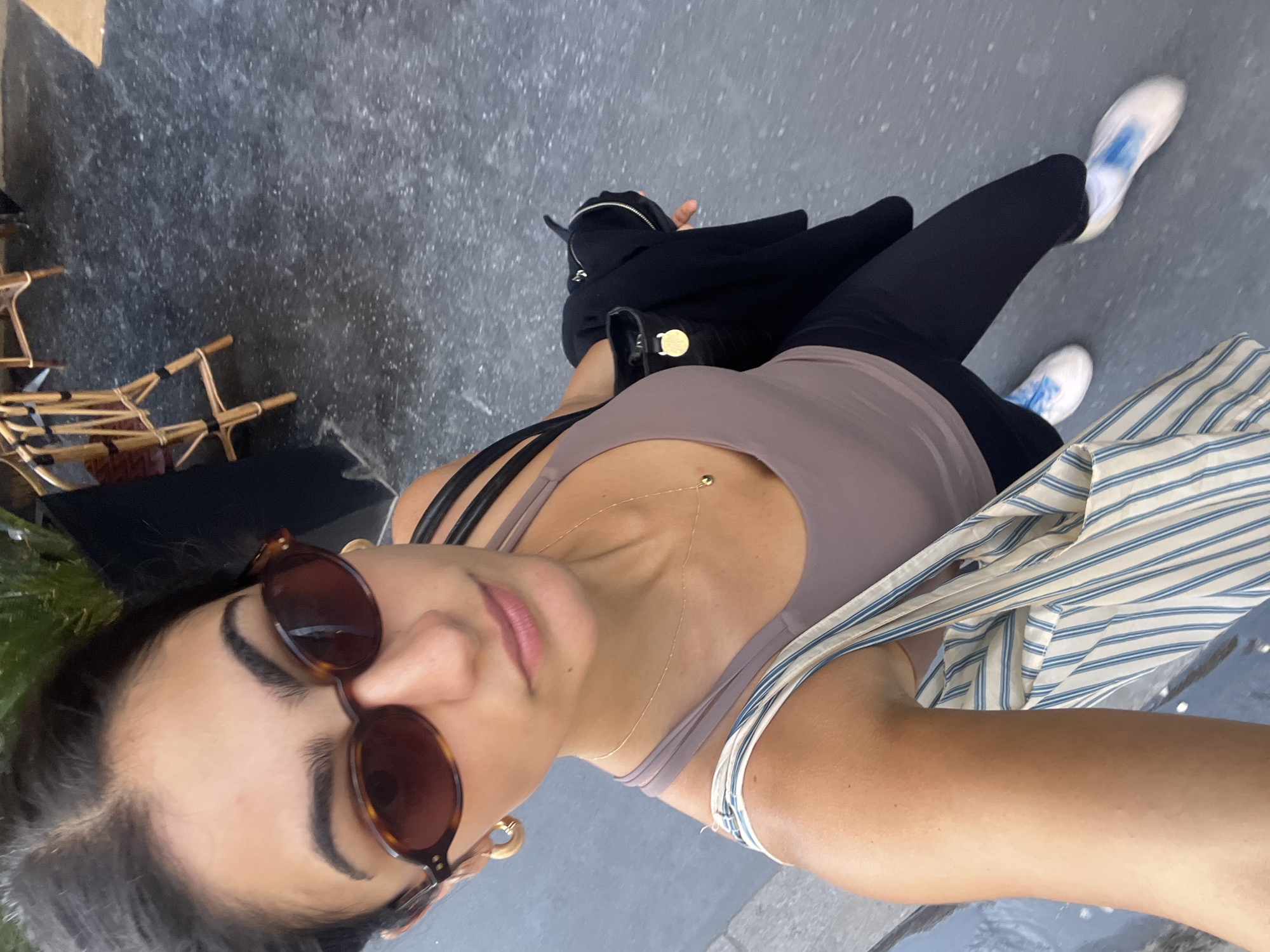 A top-down selfie captured on a street in what appears to be Paris, France, shows a woman from her head to her feet. The woman is the primary subject, seen looking directly at the camera with a neutral expression. Her dark hair is pulled back, and she wears large dark brown sunglasses that obscure her eyes. She has defined eyebrows and her lips are slightly parted.
She is dressed in a casual outfit consisting of a mauve or taupe-colored tank top or sports bra. A delicate gold chain necklace with a small circular pendant hangs around her neck, and a small belly button piercing is visible. A light blue and white striped long-sleeved shirt is tied around her waist. She wears black leggings and white sneakers with light blue accents or laces. A black shoulder bag with a gold clasp is visible on her side, resting near her hip. Gold hoop earrings with a light-colored inlay are also visible.
The setting is an outdoor urban environment, likely a sidewalk or street, characterized by dark grey, speckled paving. To the left of the frame, several traditional wicker or rattan cafe chairs are partially visible, suggesting the presence of a cafe, which is a common sight in Paris. Bright, natural light indicates it is daytime, likely sunny. No discernible text is visible on any objects or in the background.