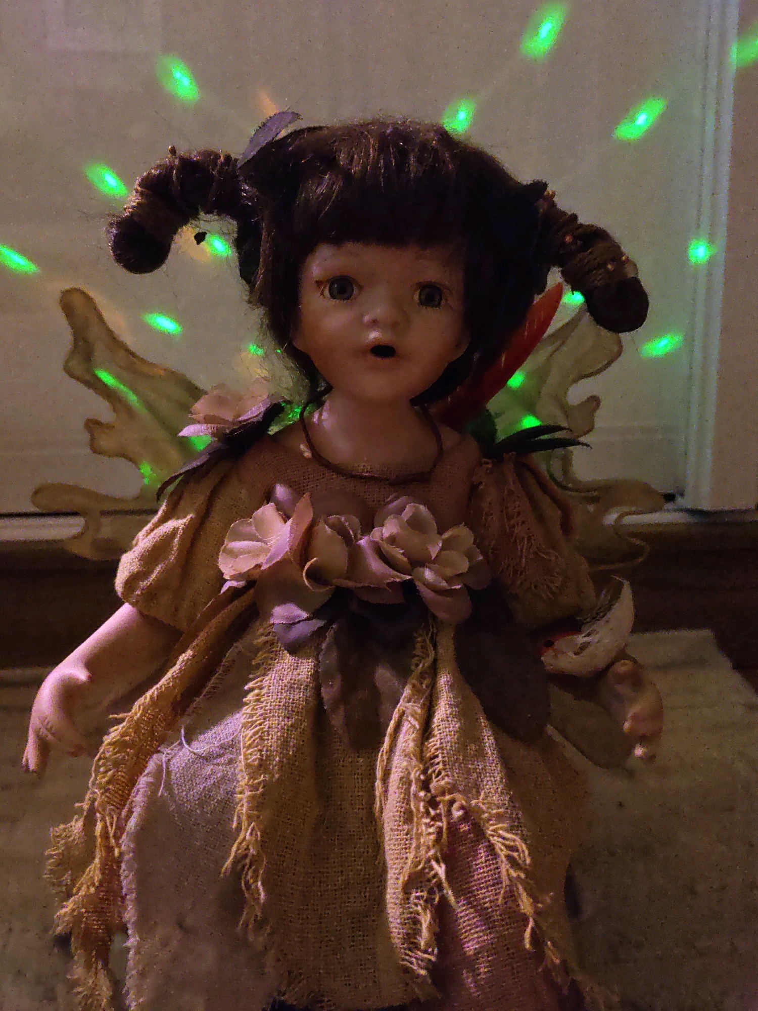 The image shows a doll resembling a fairy or sprite, positioned centrally. The doll has dark brown hair styled into two large, coiled pigtails. Its face is pale with wide, surprised eyes, an open mouth, and rosy cheeks. It is wearing a brown, burlap-like dress adorned with fabric flowers in shades of pink and beige, and leaf-like embellishments. The doll has what appear to be large, translucent wings attached to its back, made of a light-colored, possibly papery material. The background is dimly lit, with a speckled pattern of green lights that suggest a disco ball or similar lighting effect. The doll is resting on a textured surface, possibly sand or a rough rug. There is no information in the image to suggest a specific location in Rostov-on-Don, Russia.