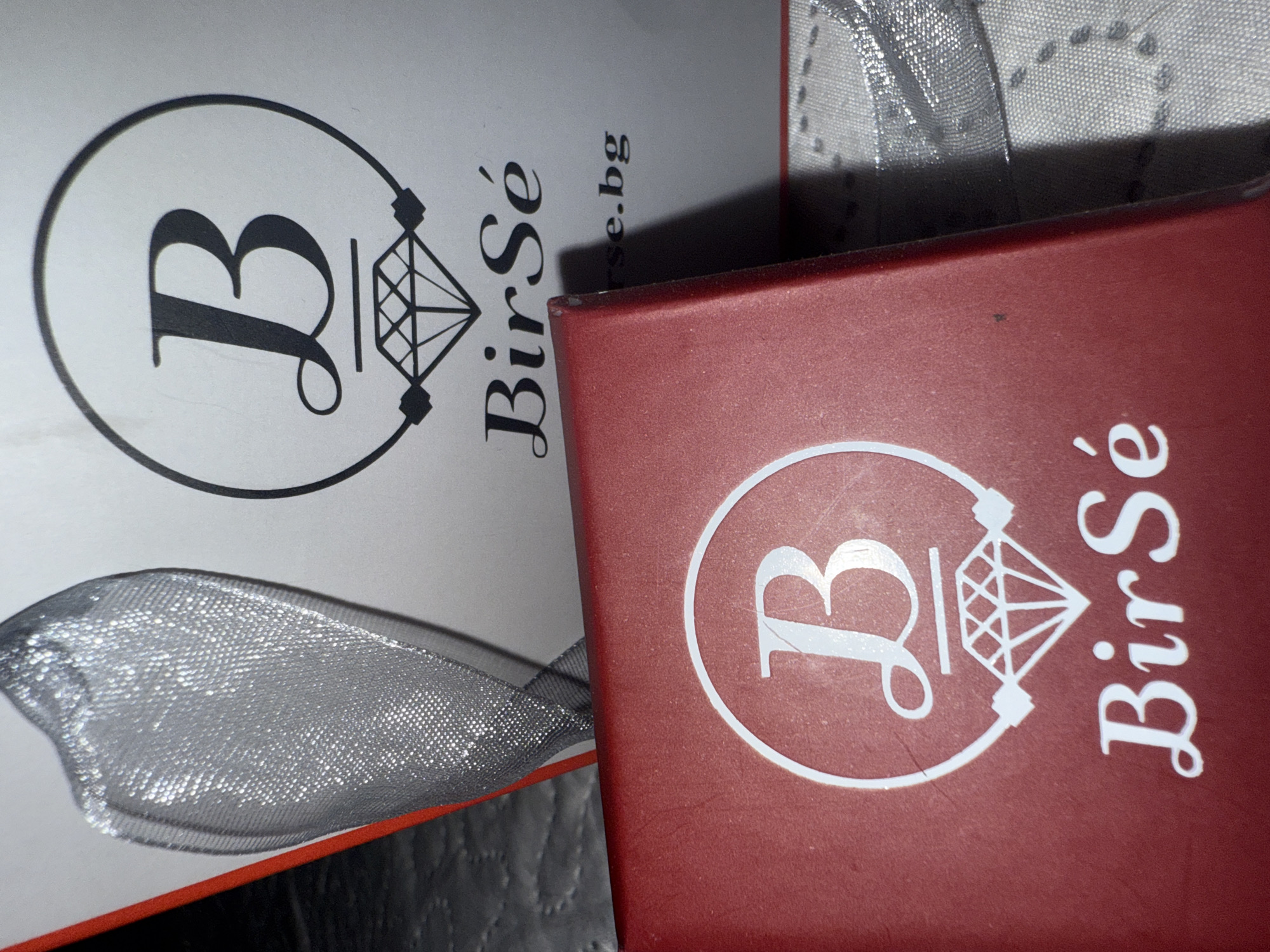 The image displays two gift boxes, one white and one red, positioned at an angle on an indoor surface. Both boxes feature a prominent logo consisting of a stylized capital letter "B" encircled, alongside a detailed illustration of a diamond. The white box's logo and text are printed in black, clearly showing "BirSé" and the website address "birse.bg". The red box showcases the identical logo and "BirSé" text in a contrasting white print.
A shimmering silver ribbon, possibly part of the packaging, is draped across the white box and extends towards the red one. The ribbon has a visible mesh texture, catching and reflecting light. The red box appears to have a matte finish with some minor scuff marks. The items are stationary, suggesting a product display or still-life arrangement. The lighting indicates an indoor setting. The "birse.bg" domain name and the provided context suggest a connection to Varna, Bulgaria.