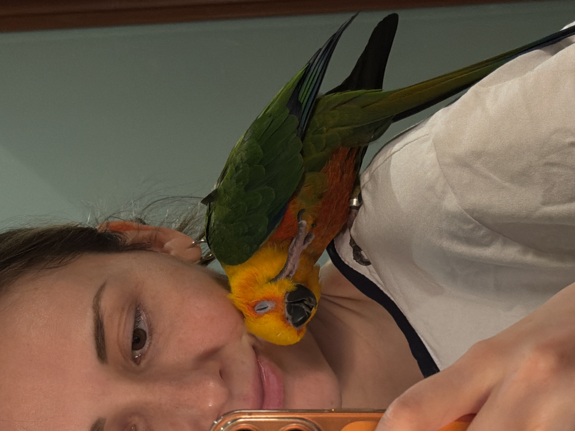 A woman is holding a cell phone and appears to be taking a selfie with a parrot perched on her shoulder. The parrot has green and yellow feathers. The woman is wearing a white shirt. The background is a light blue wall. The picture was taken indoors. No information about the city of Khimki, Russia, is available in the image.