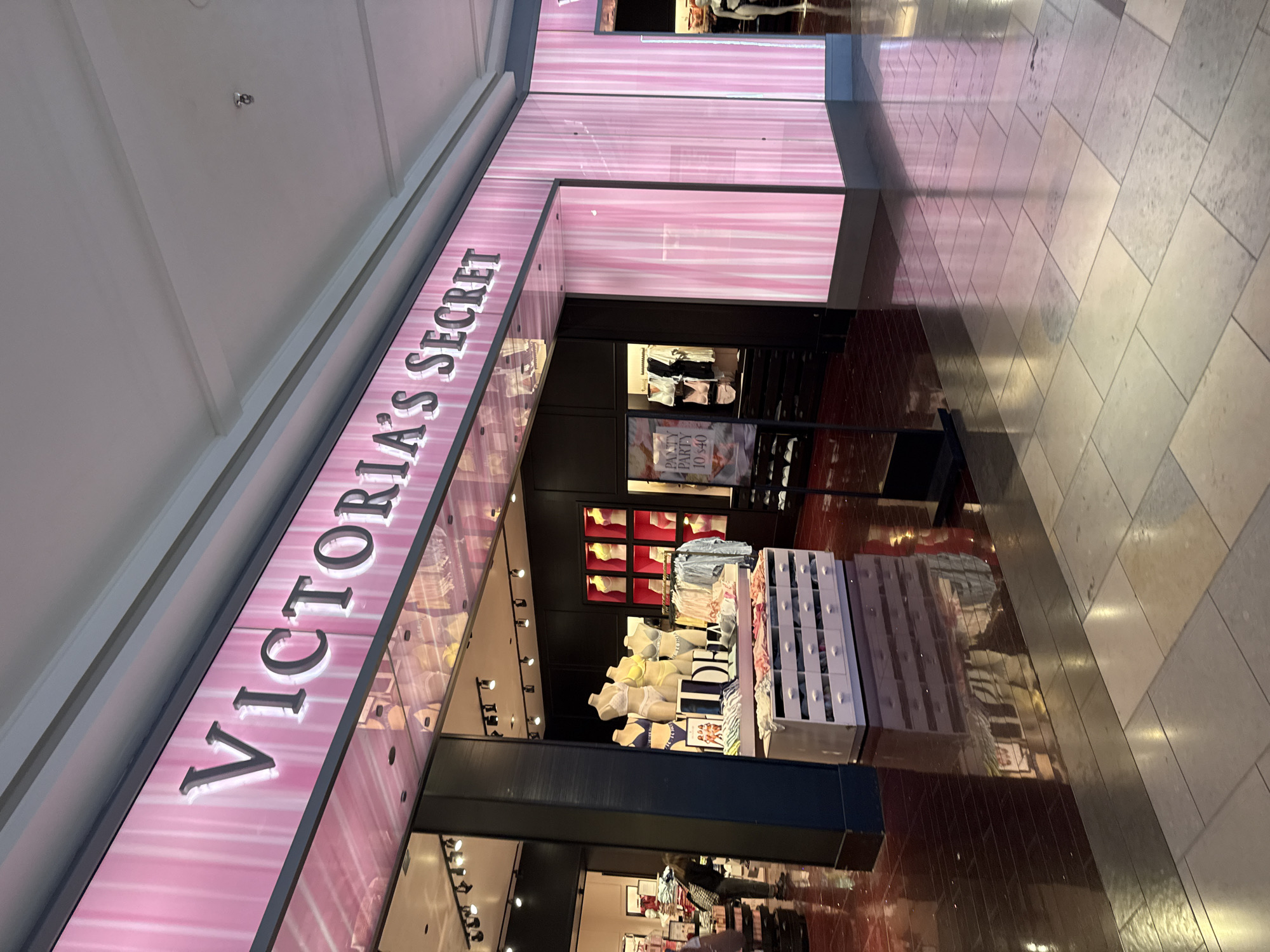 The image shows a Victoria's Secret store located inside a shopping mall. The store has a large, illuminated sign with the brand name "VICTORIA'S SECRET" in pink letters. Inside, there are displays of lingerie and other merchandise, including mannequins wearing bras and panties, along with folding tables filled with merchandise. A sign is visible advertising a "panty party" for "$10-$40". The flooring outside the store consists of large, light-colored tiles. The overall environment is brightly lit and appears to be a standard retail setting.
While the photograph does not explicitly state the location is within San Antonio, Texas, United States of America, the location of Victoria's Secret in that city could be assumed to be within a large shopping mall. The architecture and retail environment in the image are consistent with stores in such malls.