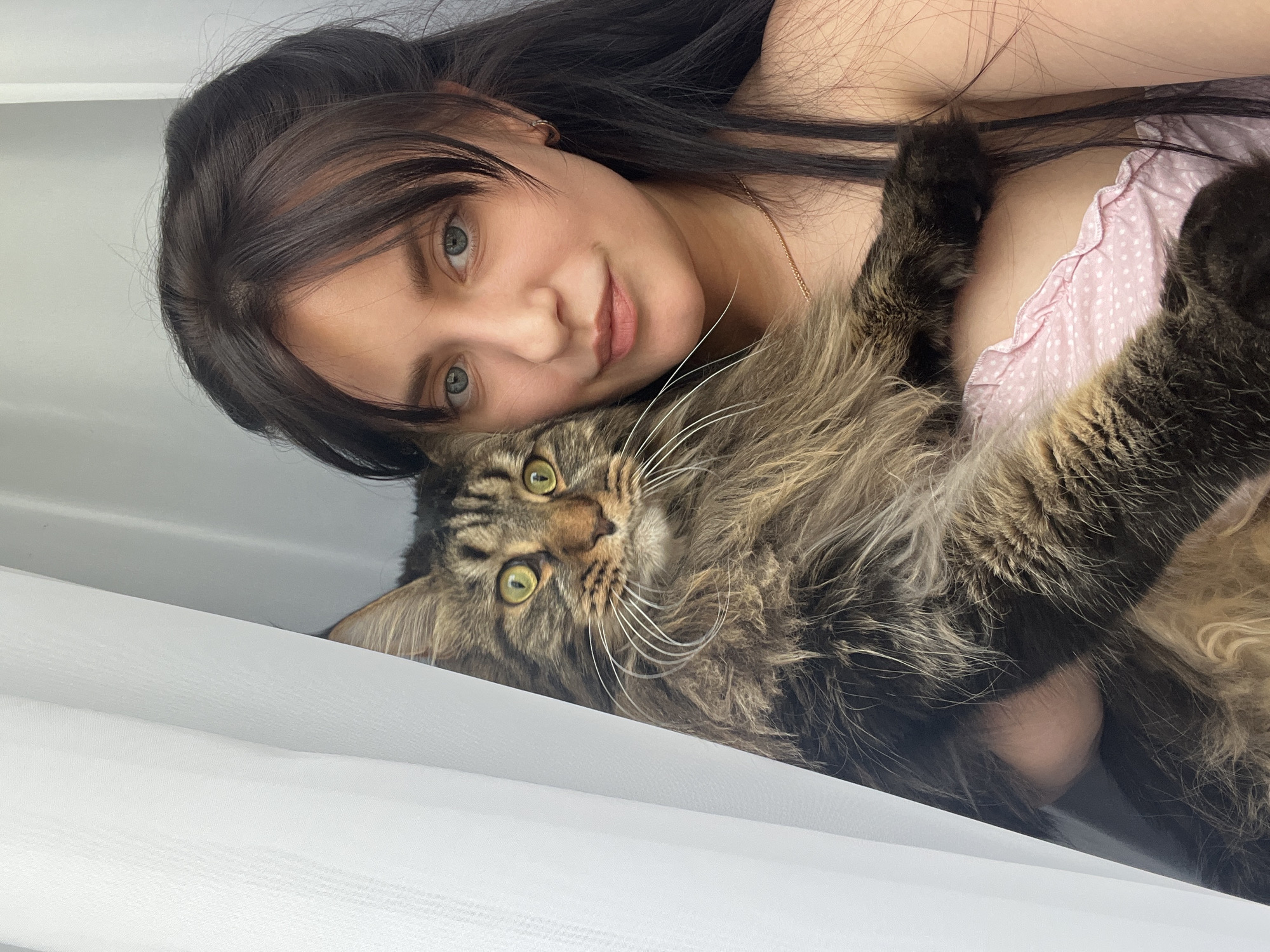 A woman with dark hair and blue eyes is holding a large, fluffy tabby cat. The woman is wearing a pink top with white polka dots. The cat is being held close to her chest and is looking towards the camera. The background is a white curtain. The image appears to have been taken indoors. There is no discernible location in the city of Марковский, Russia.