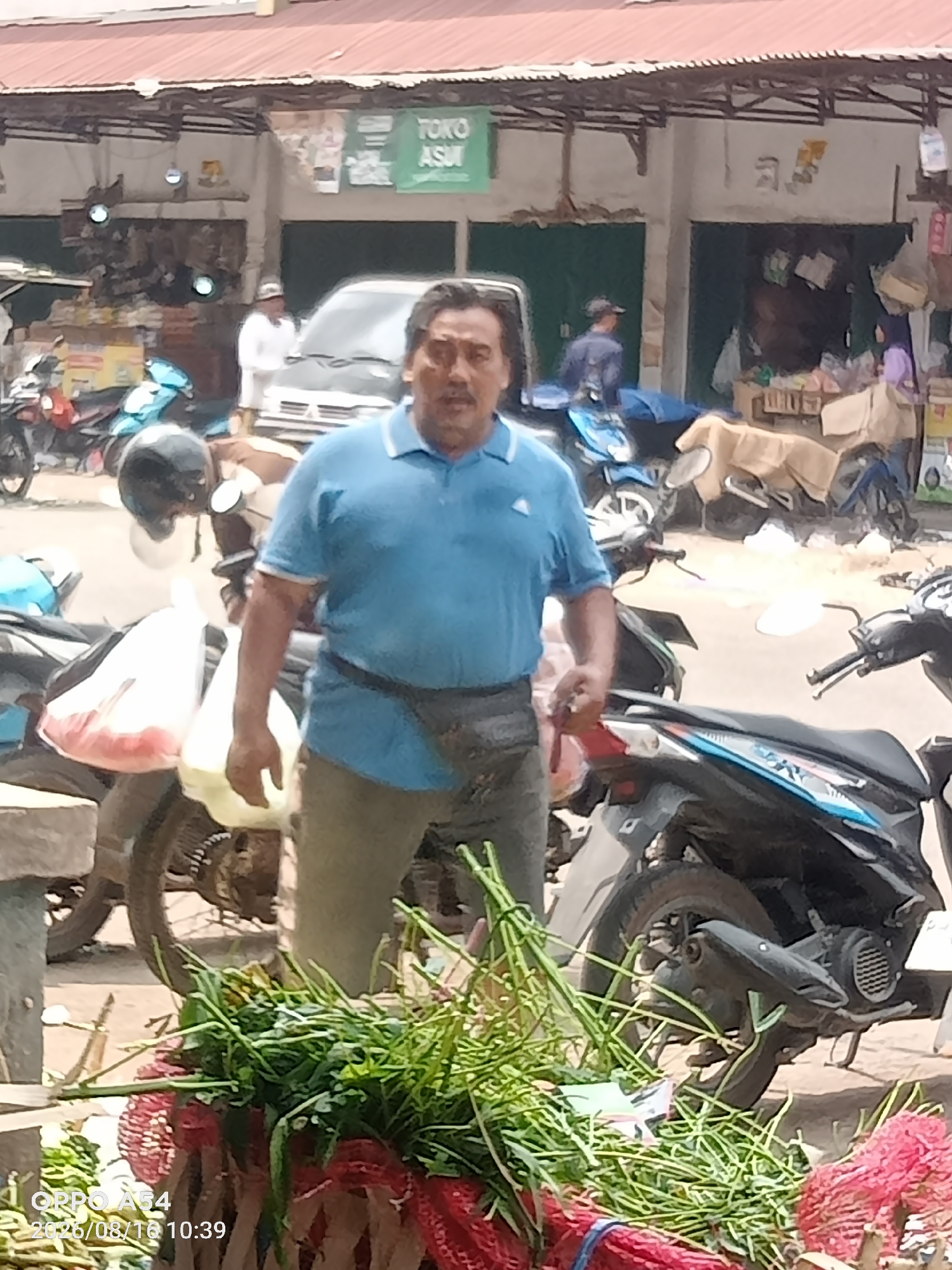 A man in a blue polo shirt and khaki pants stands in front of several motorcycles and stalls in an outdoor market. He has a dark fanny pack around his waist. Behind him, a sign reads "TOKO ASUI". The market appears to be in Jambi, Indonesia, based on the provided information. The scene is likely a busy marketplace with various vendors and customers. The overall atmosphere seems lively and bustling.