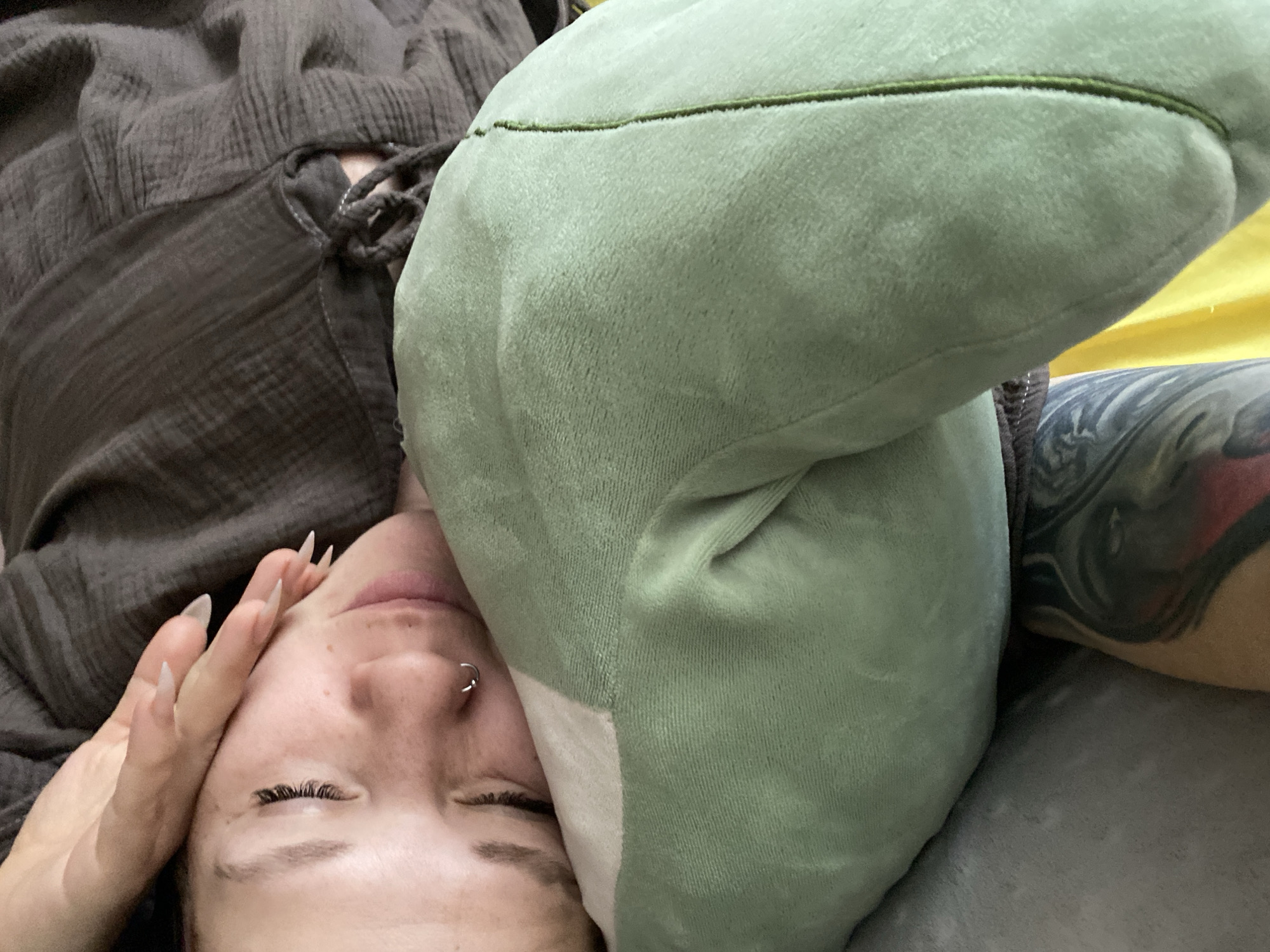 A close-up, angled shot shows a person lying down, possibly on a bed. Part of their face, including the nose (with a ring piercing), eyes closed with false eyelashes, and mouth, is visible. Their hand, with long, pointed fingernails, is near their face. They are wearing a brown, textured garment with drawstring details. A green, plush pillow obscures part of their face. A tattooed arm is visible on the right side of the frame. The setting appears to be indoors. There is no information available to identify the city of Chaikovsky, Russia as the location.