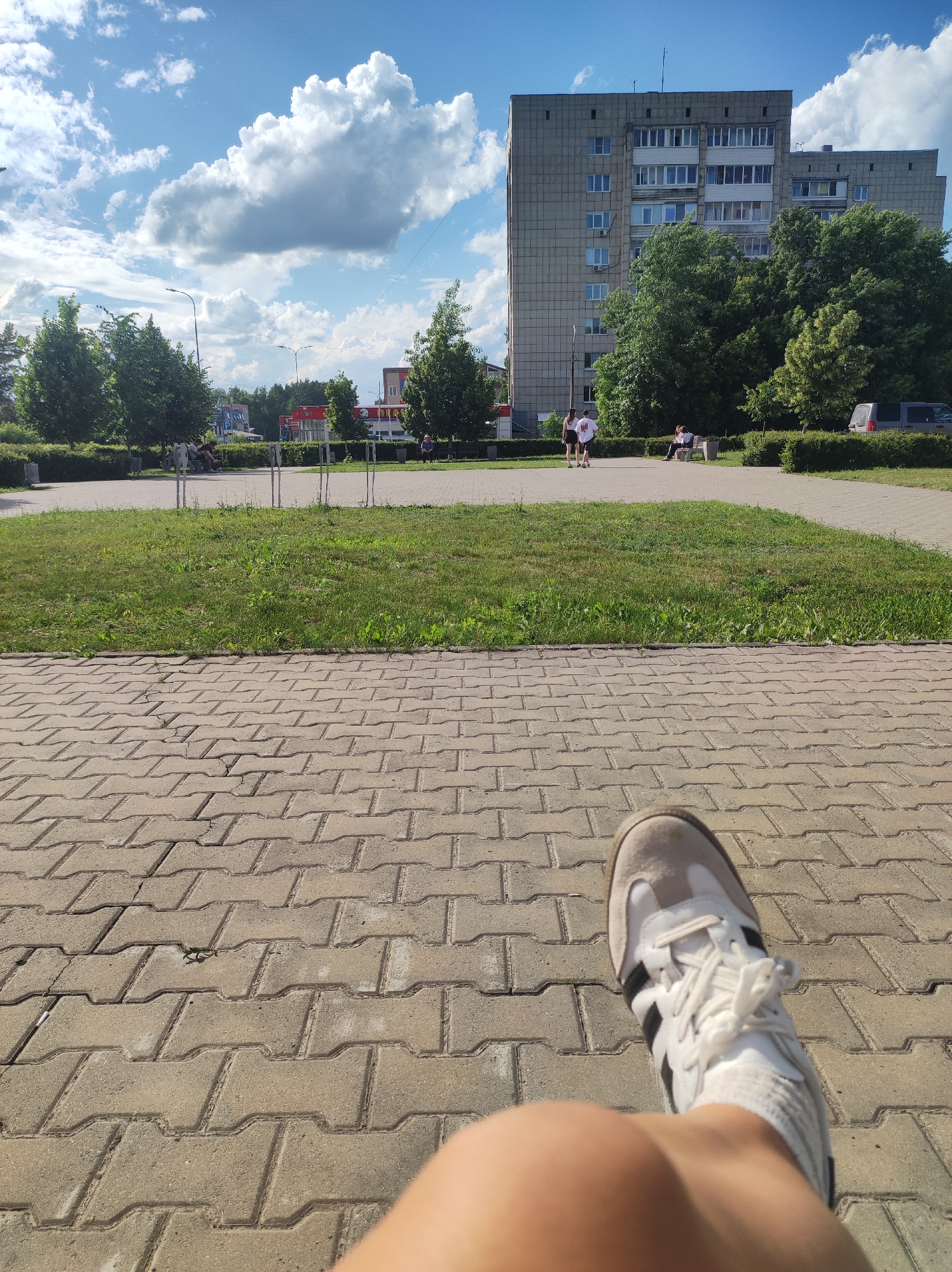 The image shows a first-person perspective of someone sitting on a paved walkway. The walkway is made of interlocking grey concrete pavers. In the foreground, the viewer's right leg and foot, clad in a white and grey athletic shoe with black stripes, are visible.
Beyond the walkway is a grassy area, and further back, a paved path with people walking and sitting on benches. Two individuals in white shirts and dark shorts are walking away from the camera. Another person is sitting on a bench in the middle ground. In the background, there is a large, grey, multi-story apartment building surrounded by lush green trees. A dark-colored van is parked to the right of the building. The sky is bright blue with scattered fluffy white clouds, suggesting it is daytime and the weather is fair. The overall scene appears to be a park or public square in a residential area.
No visible text is present in the image. The mood seems relaxed and summery.