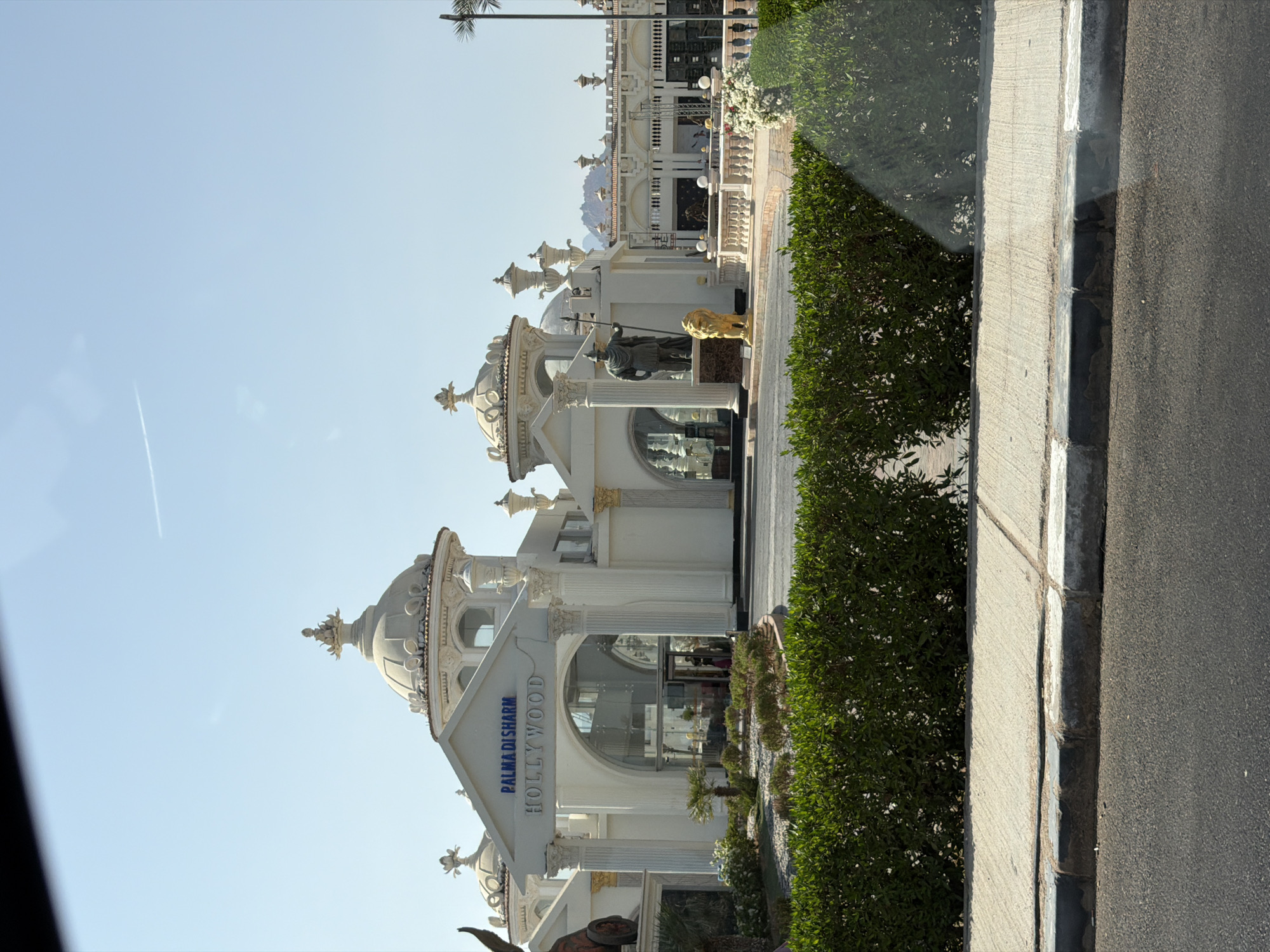 The image shows the entrance to a building with the words "PALMADI SHARM HOLLYWOOD" above the entryway. The architecture is white and ornate, with several domes and columns. There are statues of a soldier and a lion in front of the building. The building appears to be a hotel or resort. In the foreground, there is a green hedge and a sidewalk. The sky is clear and blue.