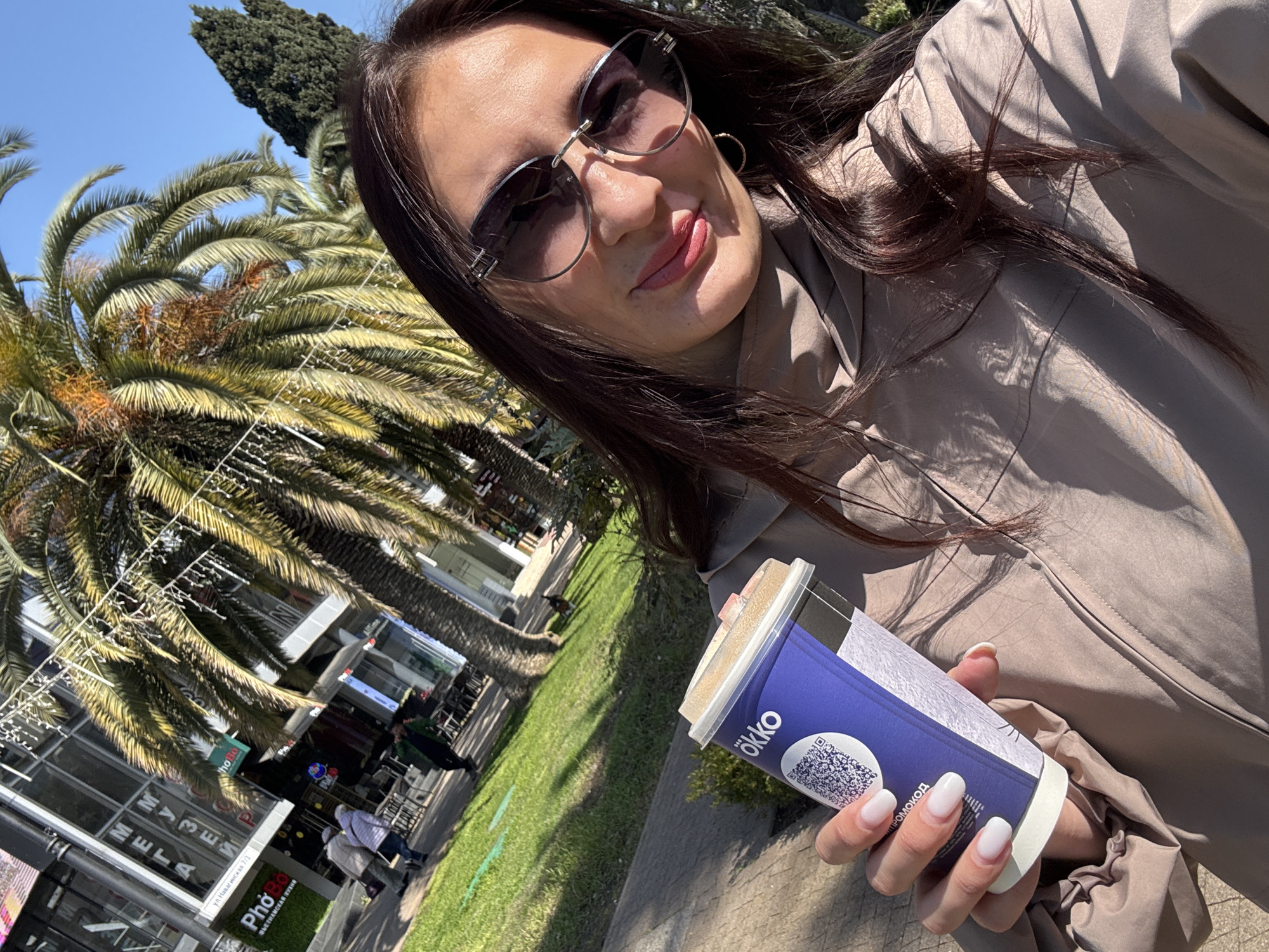 A woman in sunglasses holds a cup of coffee on a sunny day in Sochi, Russia. The cup is blue and white with the word "ökko" on it and a QR code. In the background are palm trees and a building with the signs "MY3EM" and "META". There is also a sign in Russian, "ул. Навагинская 7/3" which translates to "7/3 Navaginskaya street". The sign "Phở Bo ВЬЕТНАМСКАЯ КУХНЯ" indicates a Vietnamese restaurant. People are walking along the sidewalk.