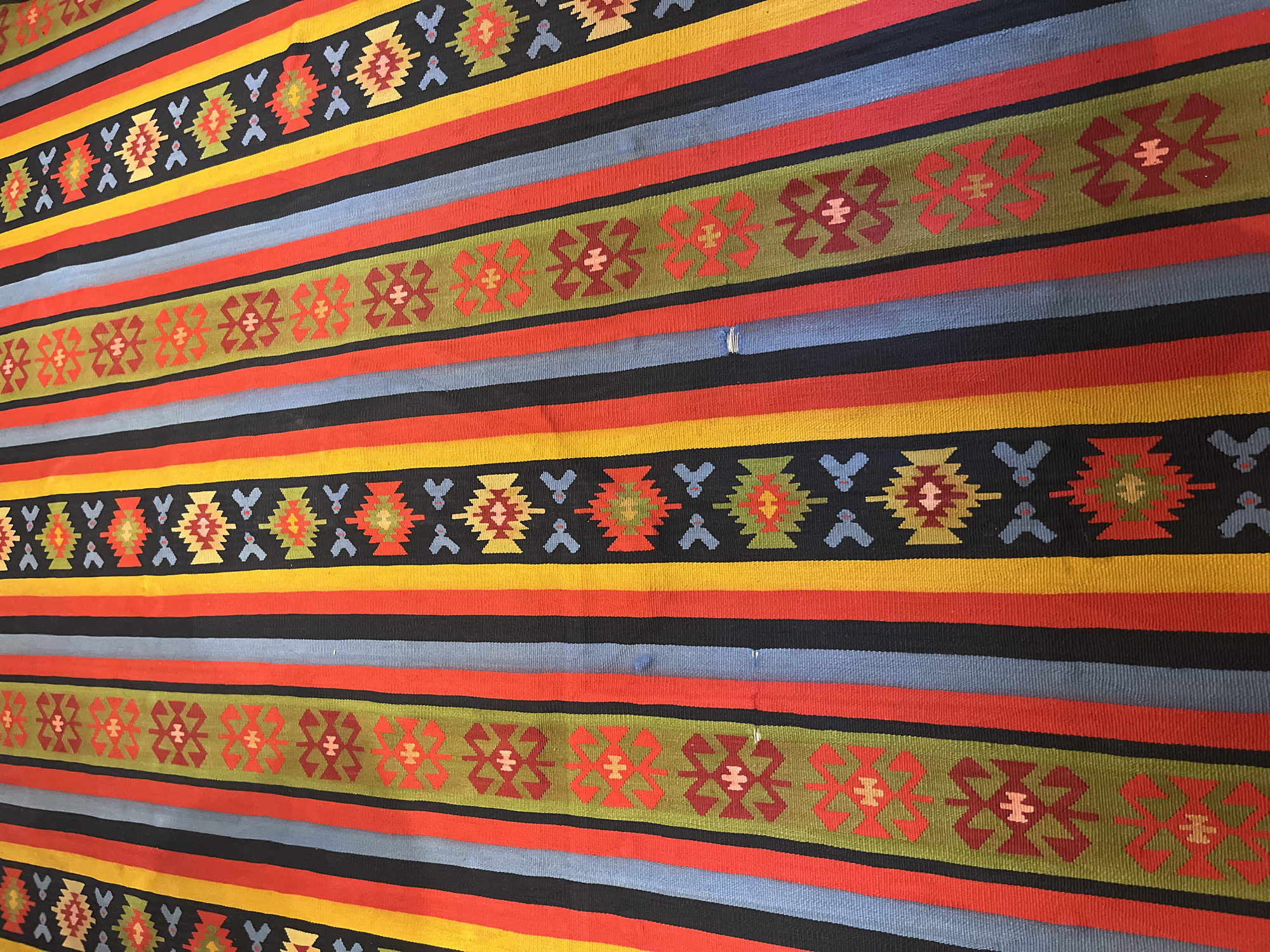 This image depicts a close-up view of a Kilim rug with a striped pattern. The rug features alternating vertical stripes of various colors, including red, black, yellow, blue, and green. Interspersed within some of the stripes are decorative geometric motifs, primarily in shades of red, green, and yellow. The rug appears to be laid out flat, with the texture of the woven material visible. The lighting suggests an indoor setting, possibly natural light. There are no people or animals present in the image. This rug is part of the collection at the Zheravna Museum in Zheravna, Bulgaria, a town known for its well-preserved 19th-century Revival architecture.