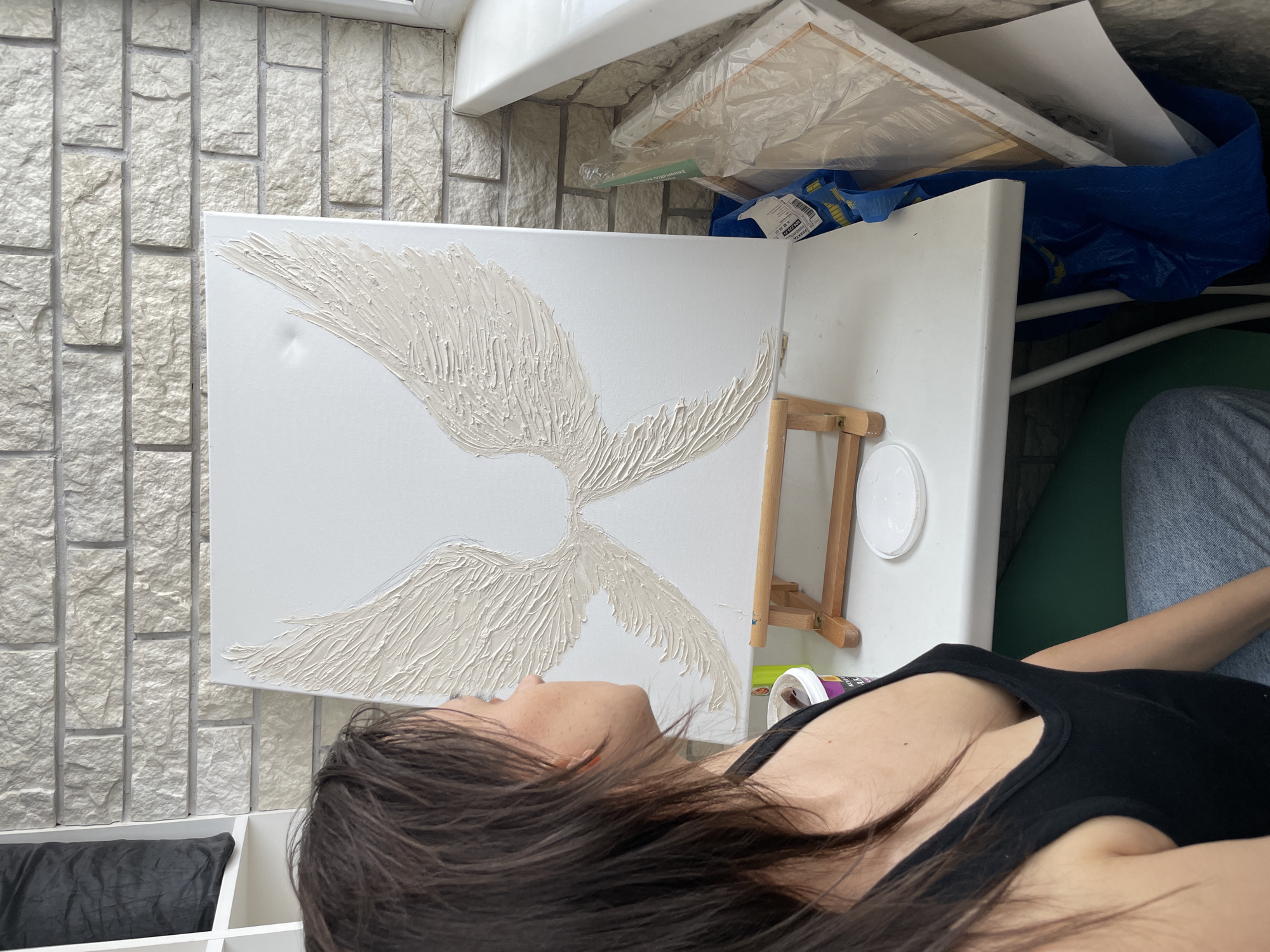 A woman with dark hair is lying down, facing upwards, near a white canvas featuring a sculpted, off-white design resembling wings. The canvas is propped up on a white surface, with a wooden easel partially visible. A white, round paint container is on the surface. Above the canvas is a collection of items, including more canvases, a blue IKEA bag, and other miscellaneous materials. The background features a wall constructed of light-colored brick-like tiles. A black bag is in a white shelf below the canvas. The image appears to be taken indoors. The location is Unknown, Unknown.