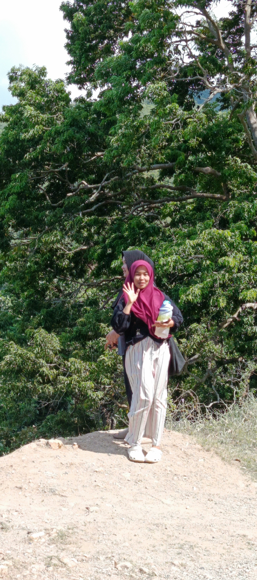 A full-shot photograph captures two women in a natural outdoor setting. The woman in the foreground, wearing a maroon hijab, a black top, and striped pants, stands facing the camera and waving. She is holding a white bottle with a blue lid. Behind her, another woman, dressed in a black hijab and dark clothing, is partially visible, looking to her left. They are positioned on a dirt path, with lush green trees and foliage forming the background. The lighting suggests it is daytime, likely under natural sunlight. The overall scene evokes a relaxed, leisurely atmosphere, possibly a tourist destination or a recreational area within Leuue, Indonesia.