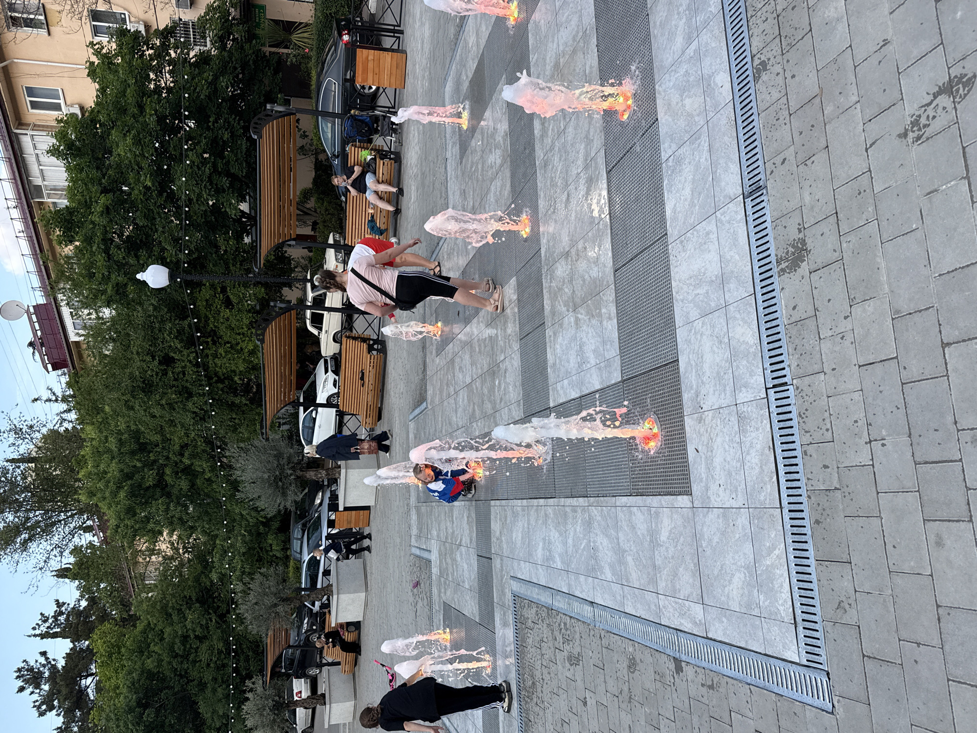 The image shows a pedestrian area in the city of Unknown, Unknown. A group of people are visible near benches in the background. In the foreground, there are water fountains where people are interacting with the water jets. The ground is paved with tiles and fitted with drainage grates. The general environment includes trees and buildings.