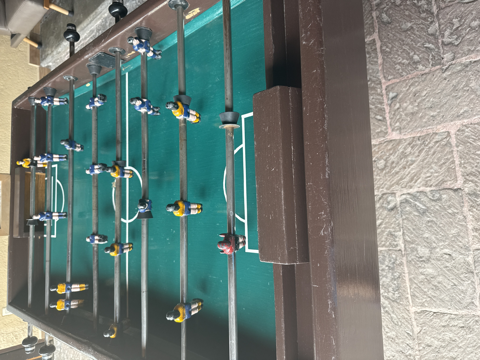 This media file captures a close-up, angled view of a vintage or well-used foosball table, set in what appears to be an indoor or covered outdoor area in Tres de Mayo, Mexico. The image is oriented vertically (rotated 90 degrees counter-clockwise), but if viewed upright, the foosball table extends from left to right.
The primary subject is the foosball table itself, which features a dark brown wooden frame showing signs of wear and tear, including scuffs and dust. The playing surface is a distinct green, marked with white lines delineating the field and goal areas. Metal rods run across the table, each equipped with several small, plastic player figurines. Most players are dressed in either blue and yellow jerseys, representing two teams, while one player figurine, positioned closer to the right goal, is notably red. Black handles are visible at the ends of some rods outside the table's frame.
No active game is taking place; the players are stationary on their rods. The lighting is diffused, suggesting an indoor setting or a shaded outdoor space, and does not indicate a specific time of day. The table rests on a textured, light-colored stone-paved floor, characterized by irregularly shaped stones and visible grout lines, hinting at traditional architecture often found in Mexico. A lighter-colored wall or structure, possibly with wooden accents, is faintly visible in the background to the left. No visible text is present in the image.