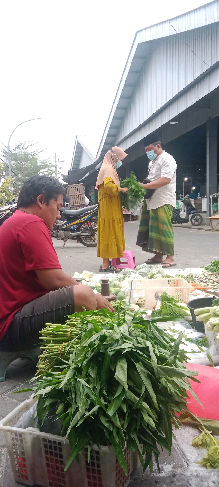 An outdoor market scene takes place in Kras, Indonesia, under an overcast sky. In the foreground, a man wearing a red t-shirt and dark shorts sits on the ground, tending to a large pile of fresh green vegetables, possibly water spinach, bundled with rubber bands in a white plastic basket. Behind him, a motorcycle is parked. In the middle ground, a woman in a yellow dress and hijab, wearing a light blue face mask, is interacting with a man in a white shirt, dark pants, and a green and white patterned sarong, also wearing a blue face mask. He holds a bunch of green vegetables. In the background, a corrugated metal roofed structure, typical of market stalls, stretches across the scene. Several crates and other produce are visible on the ground.