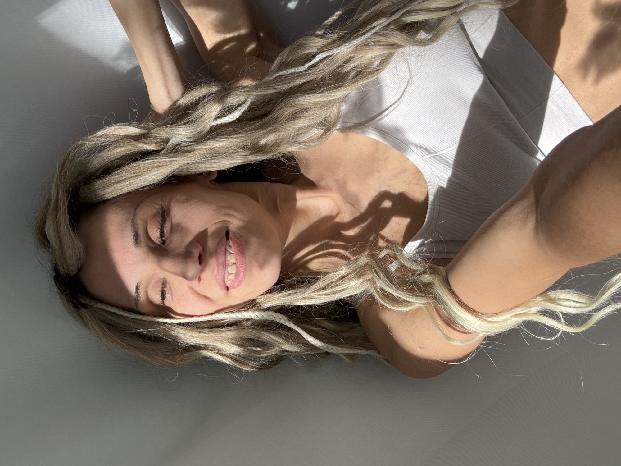 A woman with long, wavy blonde hair, interspersed with lighter braided or twisted strands, lies on her back on a light-colored surface. She is seen from an overhead perspective, looking upwards with a soft smile, her eyes partially closed in contentment. Her face is illuminated by strong, directional sunlight, casting distinct shadows across her features and the surrounding area. She wears a simple white ribbed tank top. Her arms are raised and partially visible near her head, blending into the background shadows.
The lighting suggests it is a sunny day, possibly morning or late afternoon, with direct sunlight entering an indoor space. The background is a plain, light surface, creating a minimalistic setting. The woman appears relaxed and happy, her smile conveying a sense of joy or peacefulness. No specific objects or discernible text are visible in the image. The scene takes place in Rusanovo, Russia.