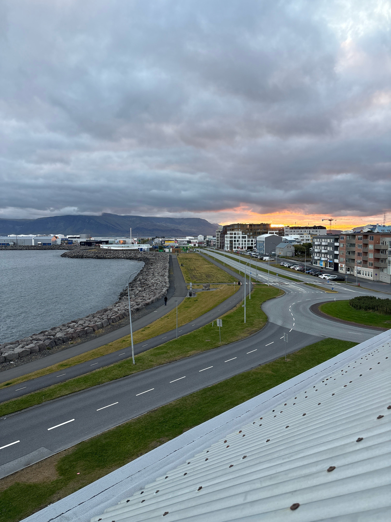The image depicts a cloudy sky over Reykjavik, Iceland. In the foreground, a white, corrugated metal roof occupies the bottom right corner. A paved road with white dashed lane markings curves through the midground, bordered by green grass. A separate paved path runs alongside the road. To the left, a rocky breakwater forms a curved edge along a body of water. Beyond the breakwater, several industrial buildings are visible, with mountains in the distance under a dramatic, overcast sky. In the background, modern buildings with clean architectural lines line the road, and cars are parked along the street. The setting sun casts a warm glow through a break in the clouds on the horizon. A single person walks along the paved path near the breakwater. A construction crane is visible in the far distance.