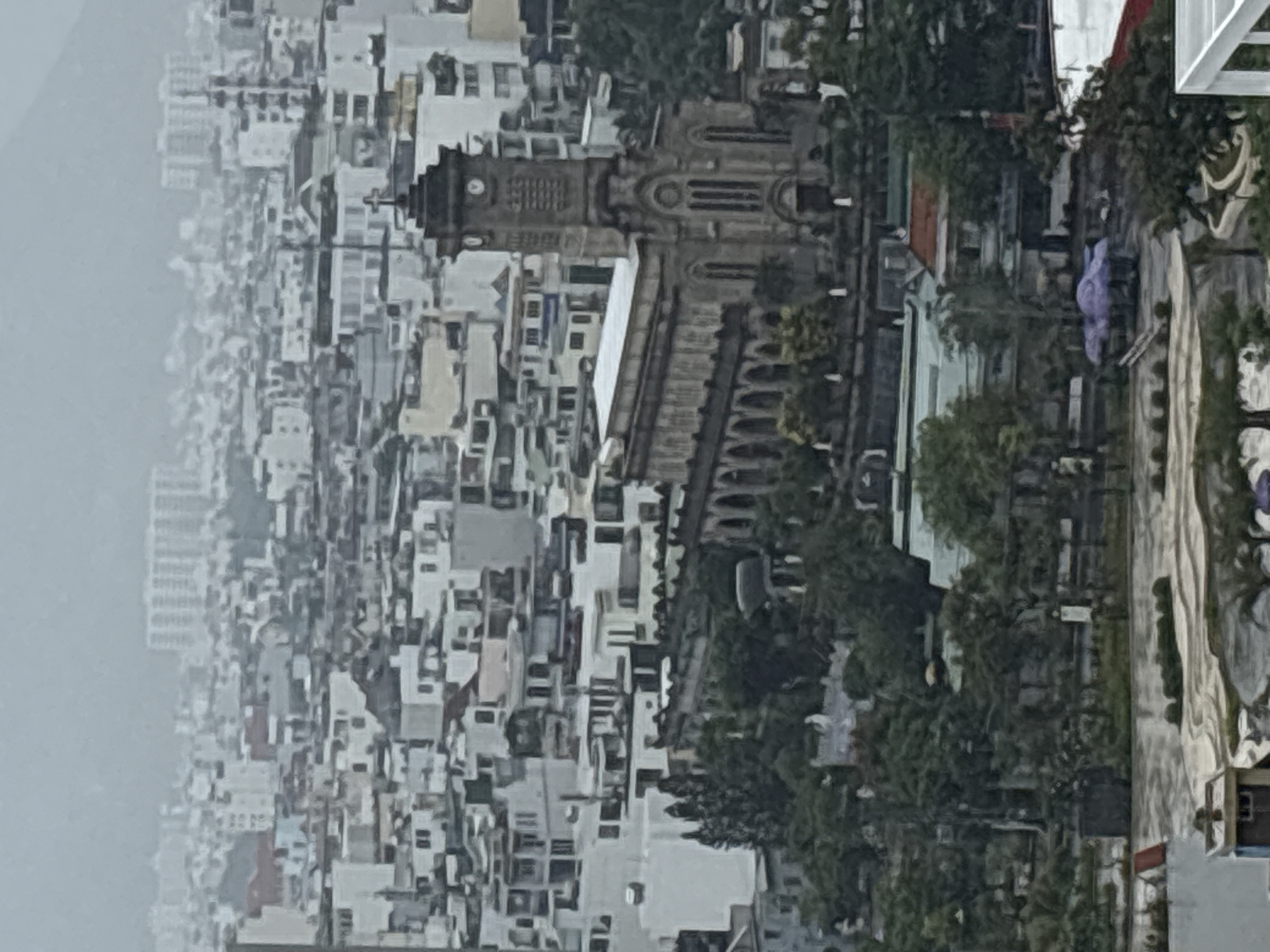 From an elevated perspective, the image captures a dense urban landscape, dominated by the Nha Trang Cathedral in Vietnam. The cathedral, a substantial stone structure with a clock tower, stands out amidst the closely packed buildings and residential structures, most of which are painted white. The left portion of the image is shrouded in mist or fog, obscuring the extent of the city. To the right of the cathedral, trees line a thoroughfare. A few figures are present, though their actions are not clearly discernible due to the distance. The scene exhibits overcast weather conditions.