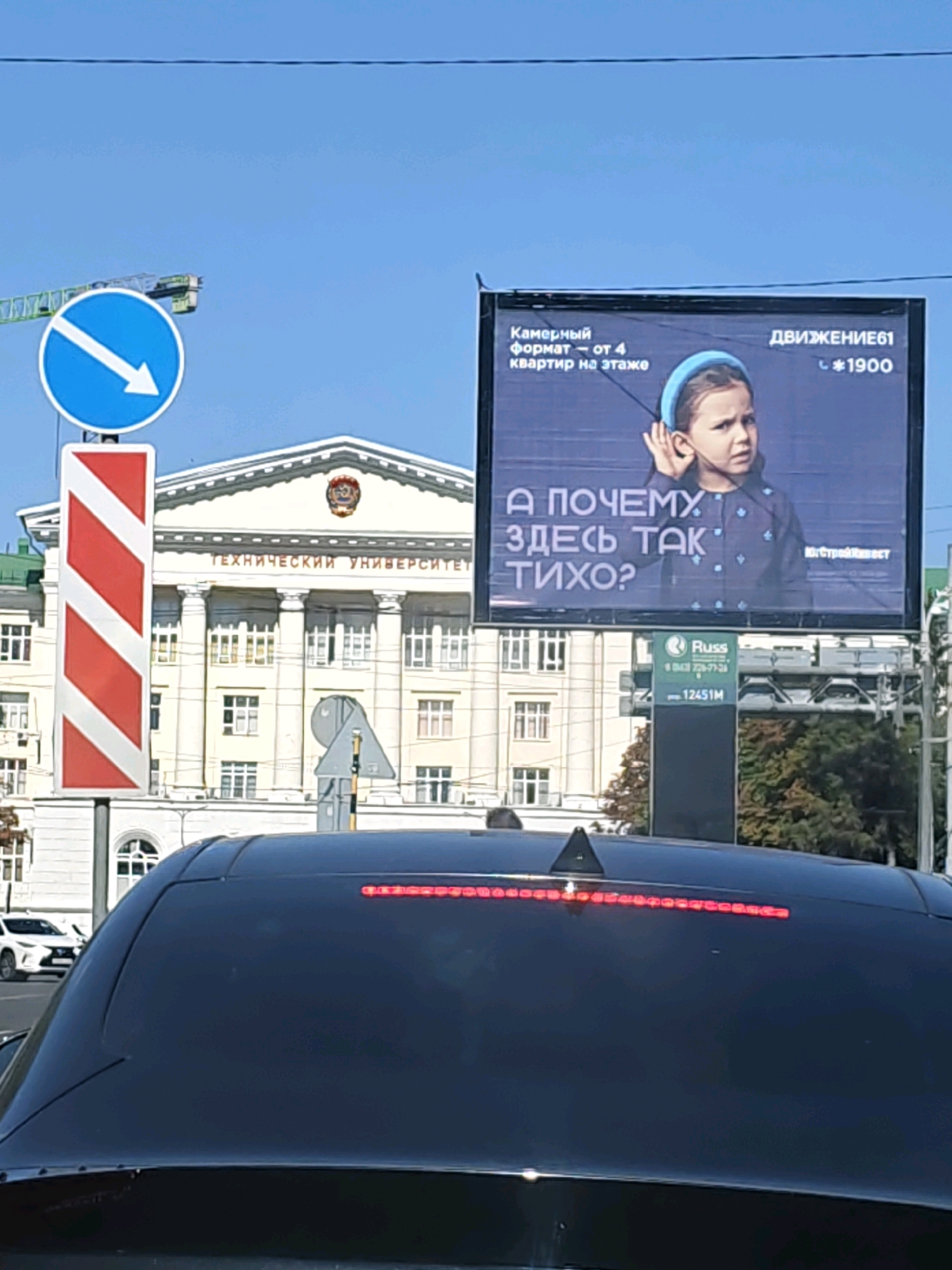 In Rostov-on-Don, Russia, a billboard displays a young girl with a questioning expression, her hand cupped to her ear. The text on the billboard reads, "Why is it so quiet here?". The billboard is positioned in front of a grand building with columns, which bears the inscription "TECHNICAL UNIVERSITY". To the left of the billboard, a blue circular road sign with a white arrow pointing right is visible, along with a red and white striped marker sign. Below the billboard, another sign shows the "Russ" logo and a phone number. The scene is captured from the perspective of a vehicle, with the rear window of a black car obscuring the lower portion of the image. The sky is clear blue, and a construction crane is visible in the distance.