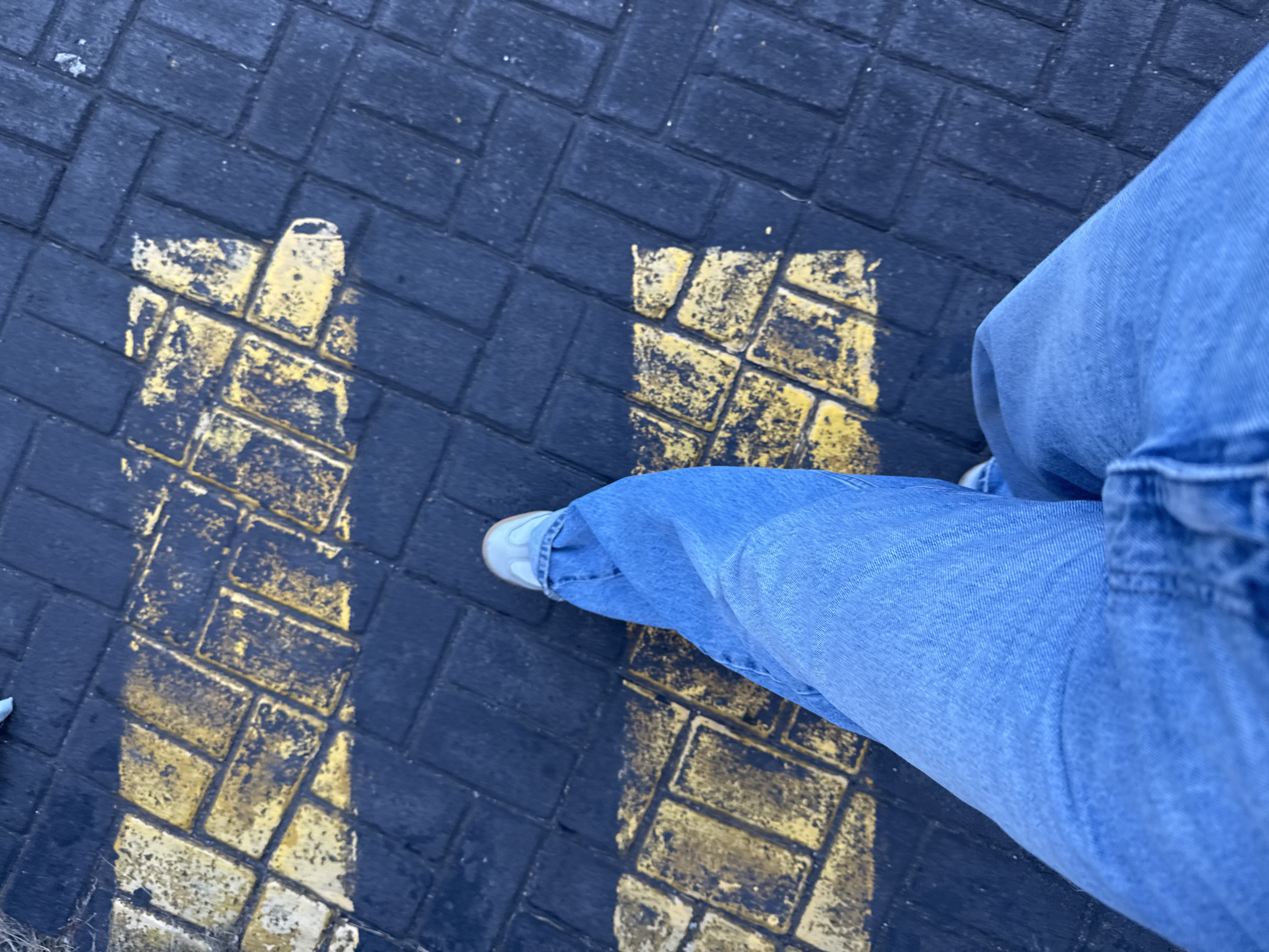 A first-person view shows the lower half of a person wearing light-blue jeans and white sneakers, standing on a dark-gray brick pavement. Painted on the pavement are two parallel lines filled with the same brick pattern in a faded yellow color, indicating a designated pedestrian walkway or a similar path. The location cannot be confirmed as Kuta, Indonesia without additional contextual information.