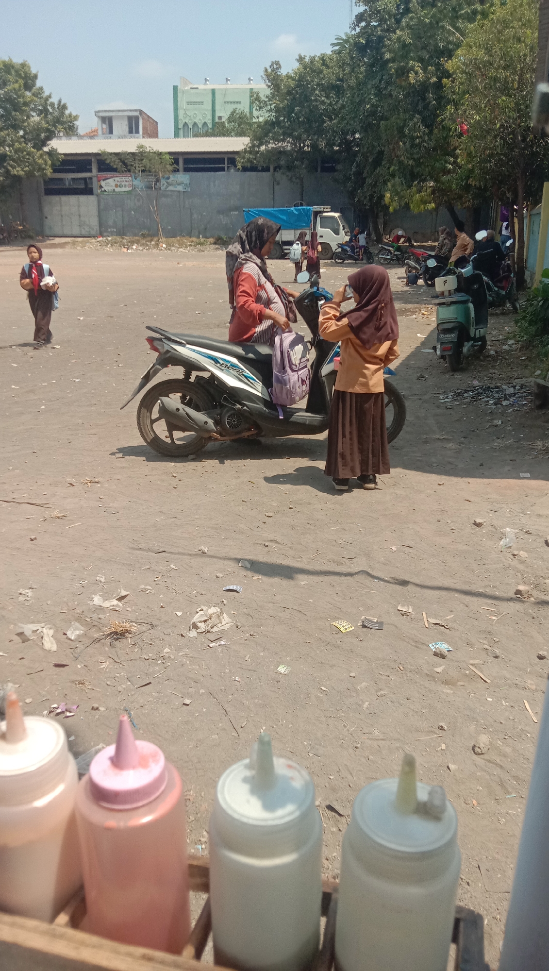 The image depicts a scene in Ngempit, Indonesia, likely near a school or educational institution given the presence of individuals in school uniforms. The foreground shows a wooden tray holding several condiment bottles, including a pink-capped one and two white ones. Beyond this, a dusty, unpaved area is visible.
In the mid-ground, two young girls in school uniforms, one wearing a brown hijab and the other a dark headscarf, are near a scooter. The girl in brown appears to be holding a phone, possibly taking a picture, while the girl with the dark headscarf is seated on the scooter and carrying a purple backpack. Another young girl in a brown uniform and a red scarf is walking away from the scooter towards the left side of the frame.
Further in the background, several people are gathered, some standing and others near parked scooters. A white truck with a blue tarp is parked behind them. Buildings with a light green facade and a white roof, possibly a mosque or other institutional building, are visible in the distance, along with trees and a concrete wall. The overall environment appears to be an outdoor, semi-urban setting with evidence of moderate litter on the ground. The sky is clear and bright, suggesting a sunny day.