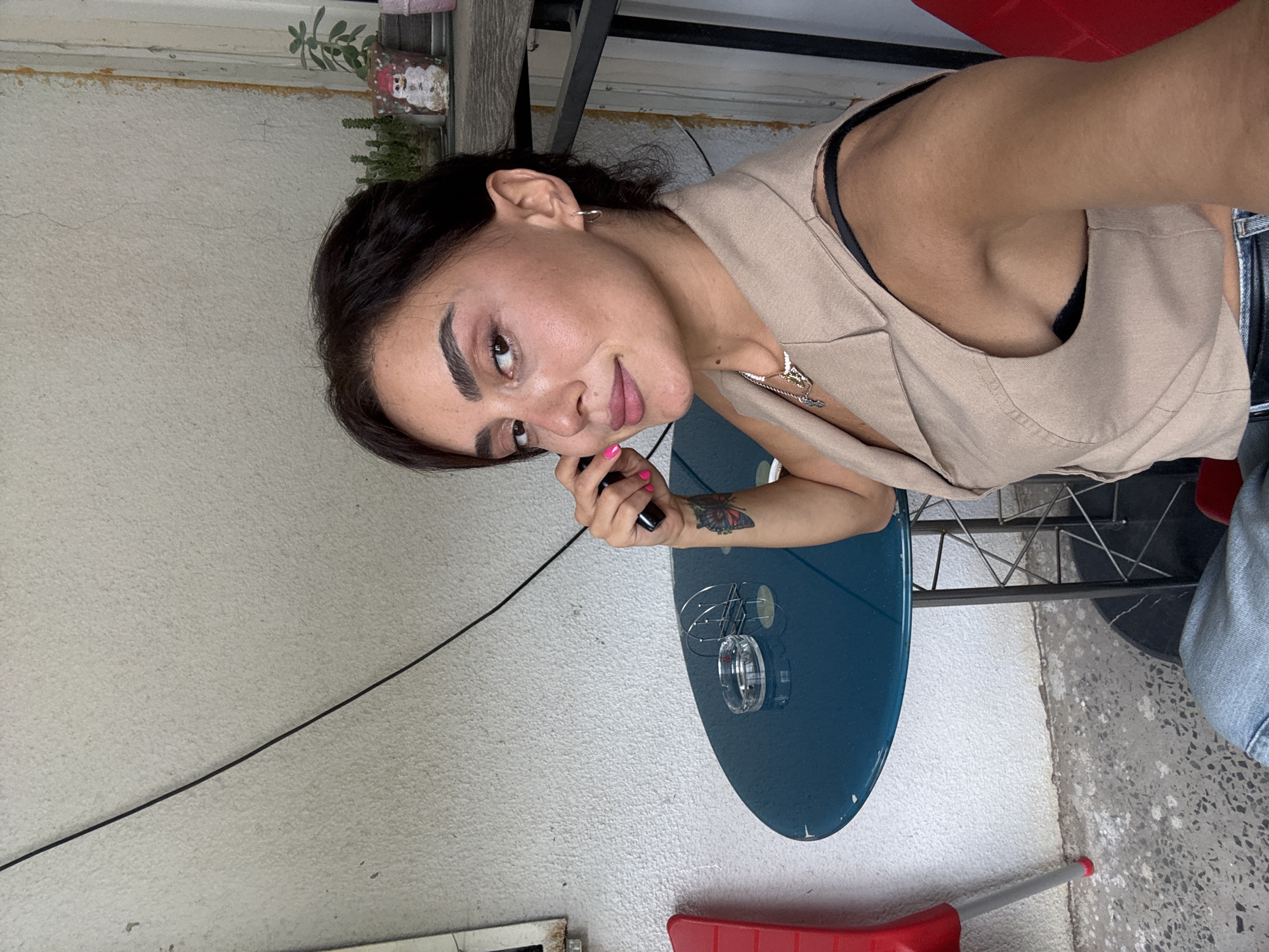 A woman with dark hair is sitting at a round, teal-colored table on a patio. She is wearing a beige vest over a black bra and has a tattoo on her left forearm. A cigarette ashtray and a napkin holder are on the table. In the background, there are red chairs, a textured white wall, and plants on a shelf. The scene appears to be outdoors, likely in a cafe or restaurant setting, possibly in Tbilisi, Georgia, given the architectural style and vegetation.