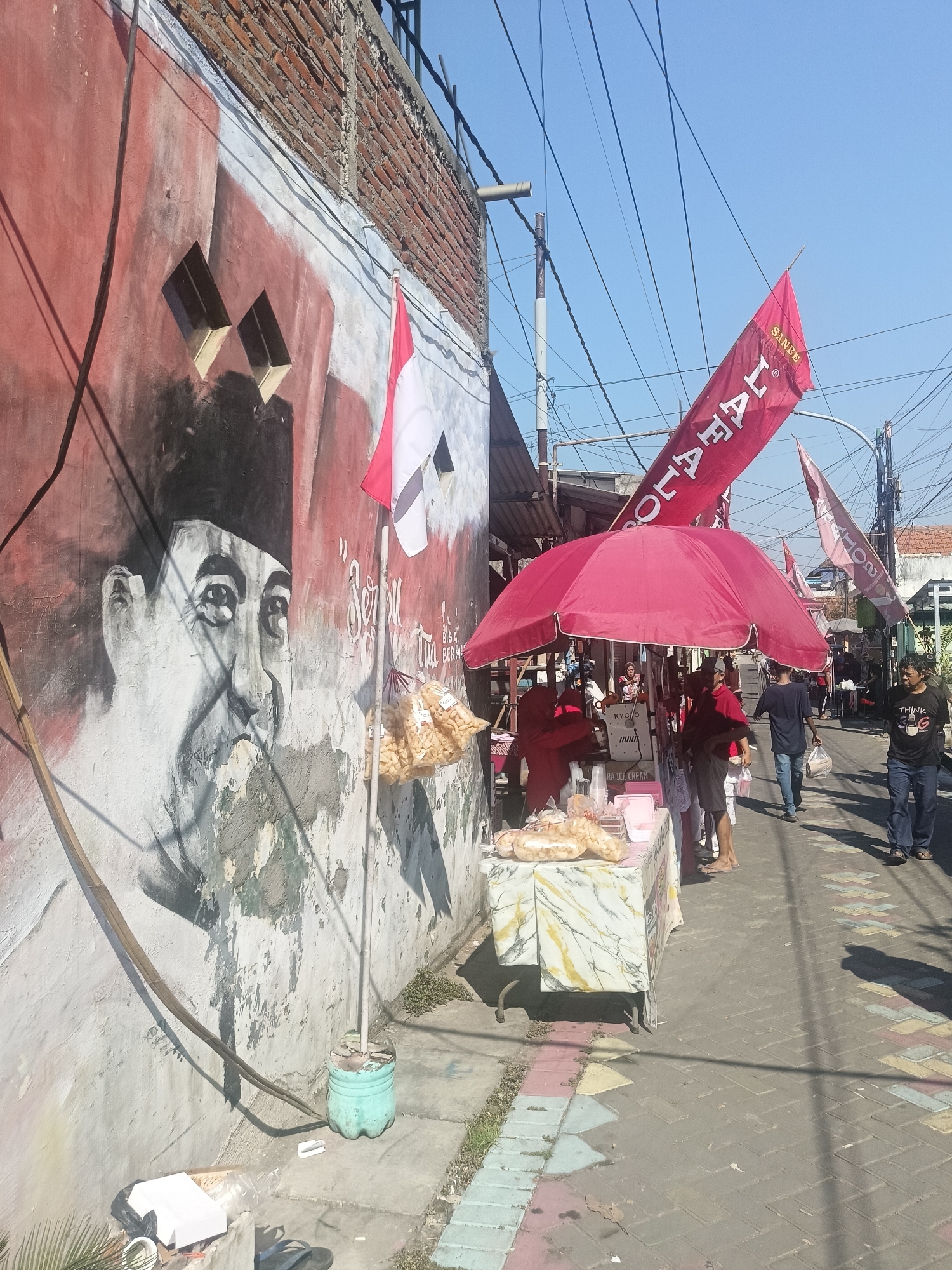 The image captures a street scene in Gedangan, Indonesia, under a clear blue sky. On the left, a rough brick wall features a large mural of a man's face, rendered in shades of white, gray, and red. The mural depicts the man wearing a dark cap and looking towards the viewer. Adjacent to the mural, a white and red flag, likely the Indonesian flag, is attached to a pole. Bags of snacks are hanging from the pole.
In the center and right of the image, a street market is set up. A large red umbrella provides shade over a stall where several people are gathered. Tables are laden with goods, including bags of snacks and pink containers. The ground is paved with patterned bricks, and a sidewalk runs alongside the street. Several individuals are walking down the street, some carrying bags, suggesting ongoing commerce. Red banners with white text are visible hanging above the market stalls. The overall atmosphere appears to be one of a bustling local market, possibly during a daytime event or celebration, given the flags and banners.